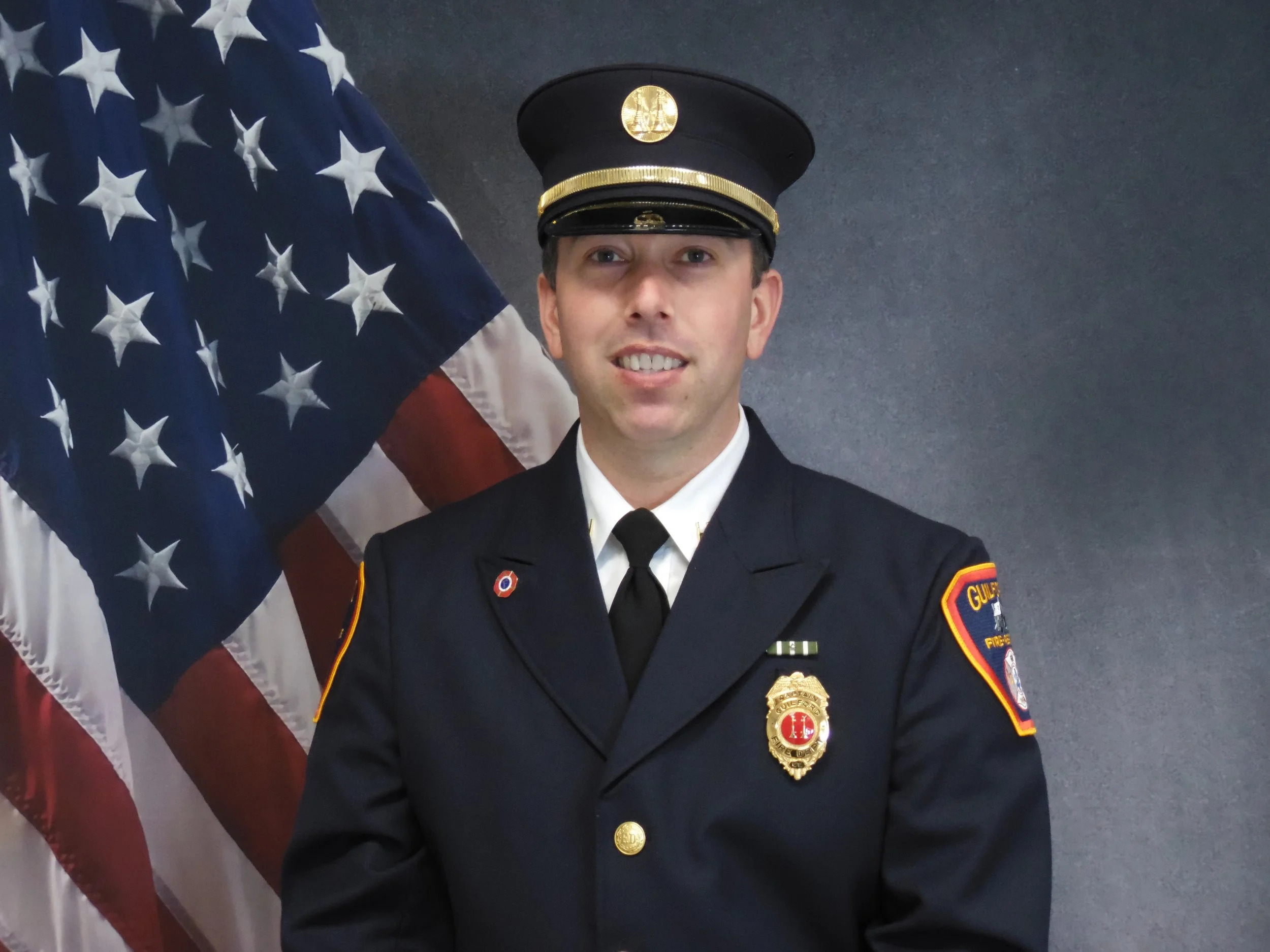 A firefighter in uniform standing in front of an American flag.