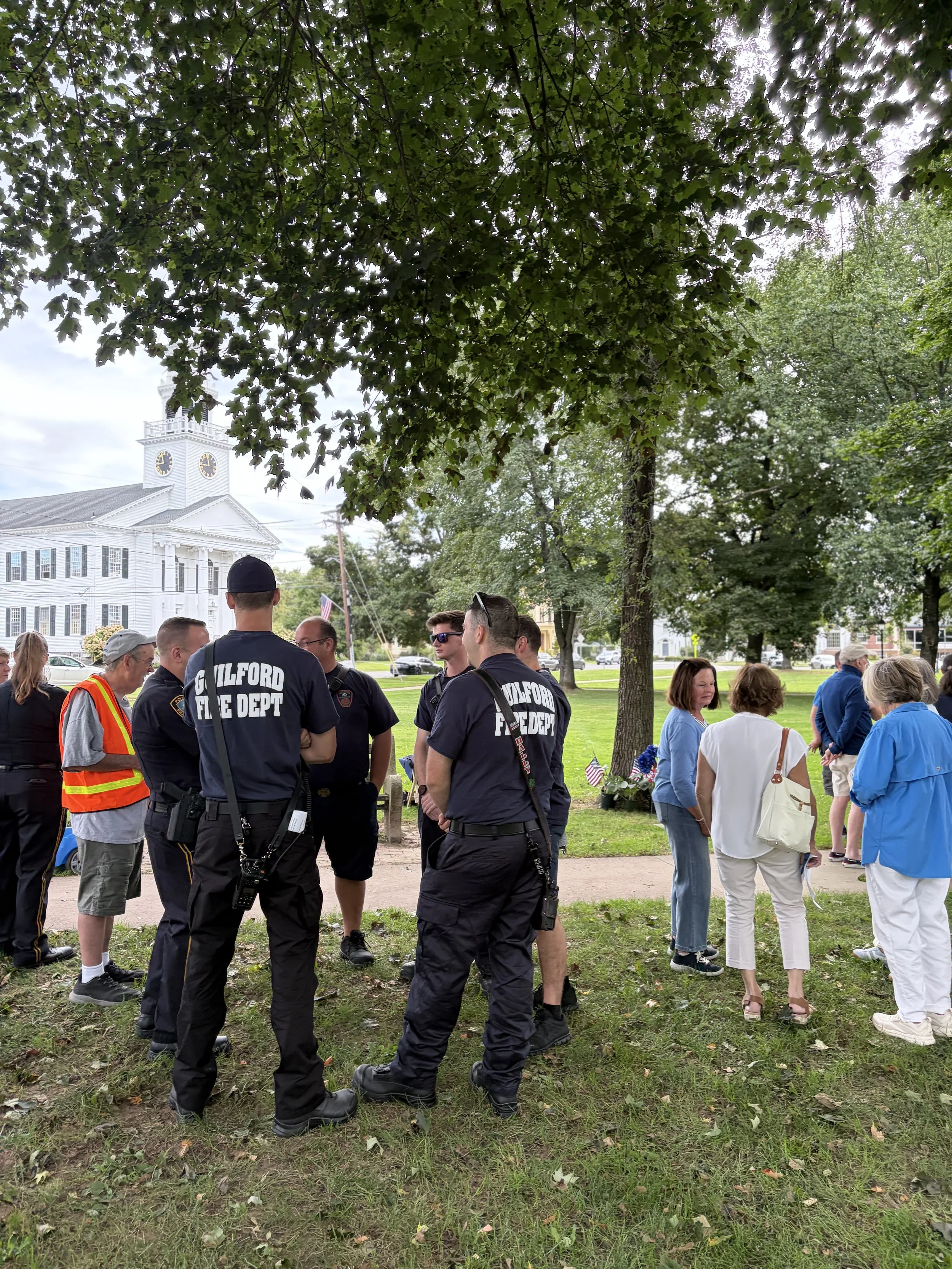 Group of people, including firefighters in uniform and civilians, gathered outdoors under trees in front of a historic white building with a clock tower.