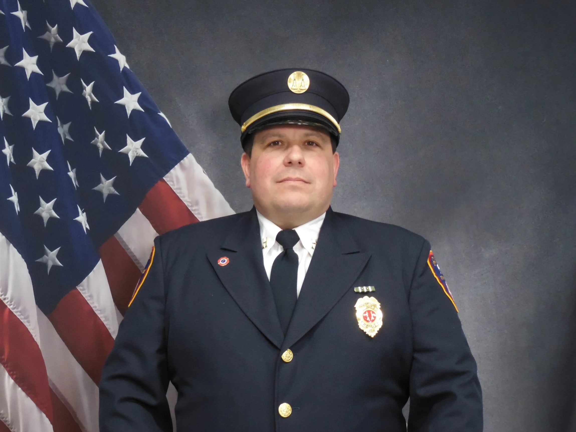 A male firefighter in a formal dress uniform with a peaked cap standing in front of an American flag and a dark background.