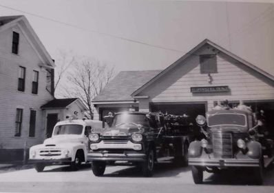 Black and white photo of two vintage trucks and a car parked on a street in front of houses.