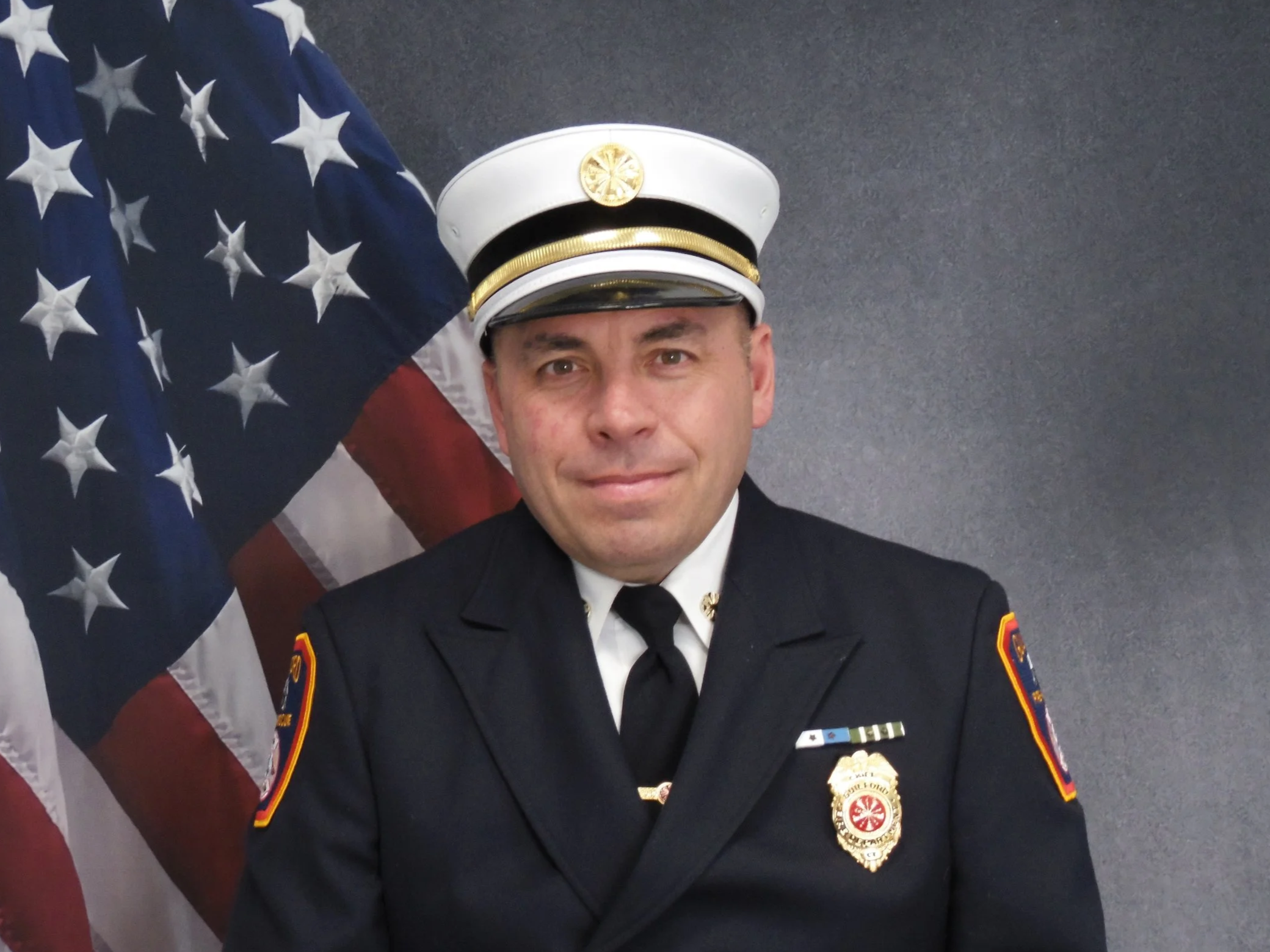 A man in a police uniform standing in front of an American flag, smiling at the camera.