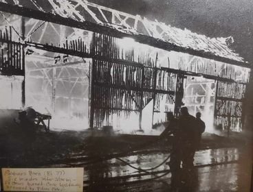 Black and white photograph of a large burning building with smoke and flames visible through the windows. Two people are in front of the building, possibly firefighters.