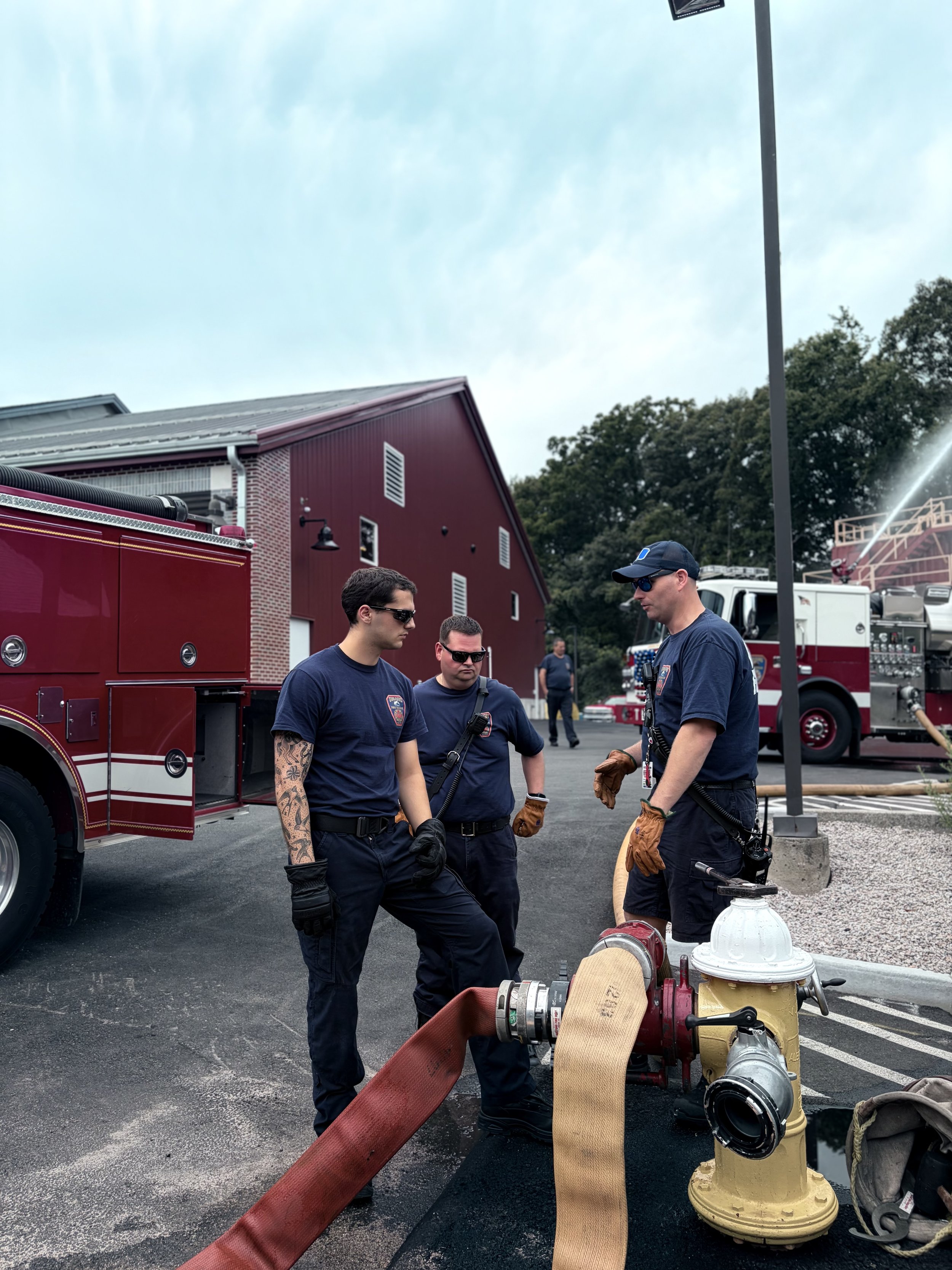 Three firefighters in navy uniforms and sunglasses standing outdoors near fire equipment, with a fire truck and building in the background.