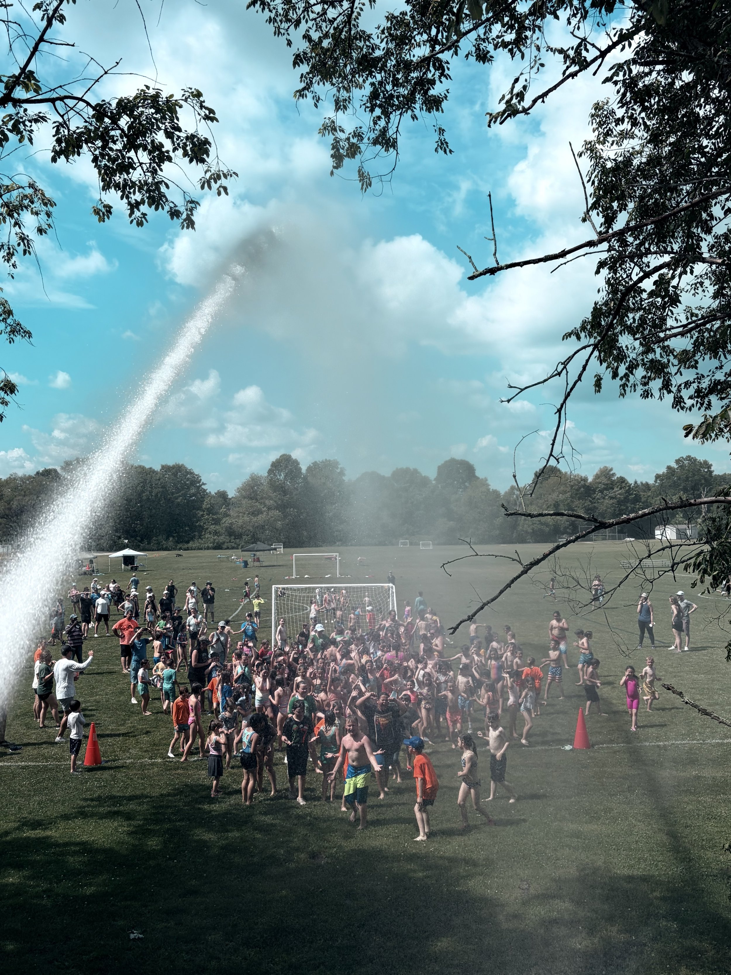 A large group of children and adults at a park participating in a water fight under a blue sky with clouds, trees, and a fountain spraying water.