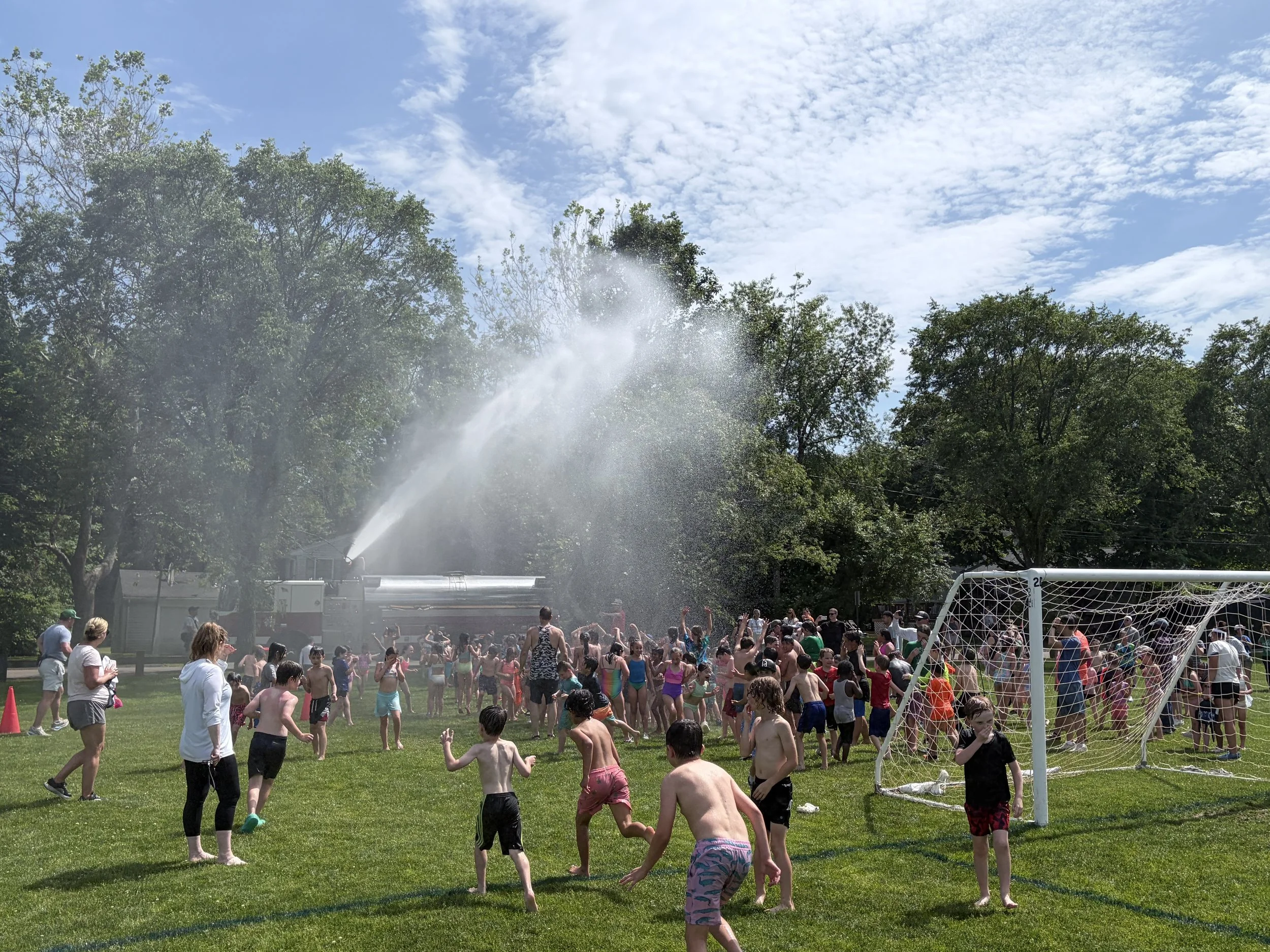 Children and adults playing in a park warmth, some wearing swimsuits, with a water spray from a fire truck and a soccer goal in the foreground.