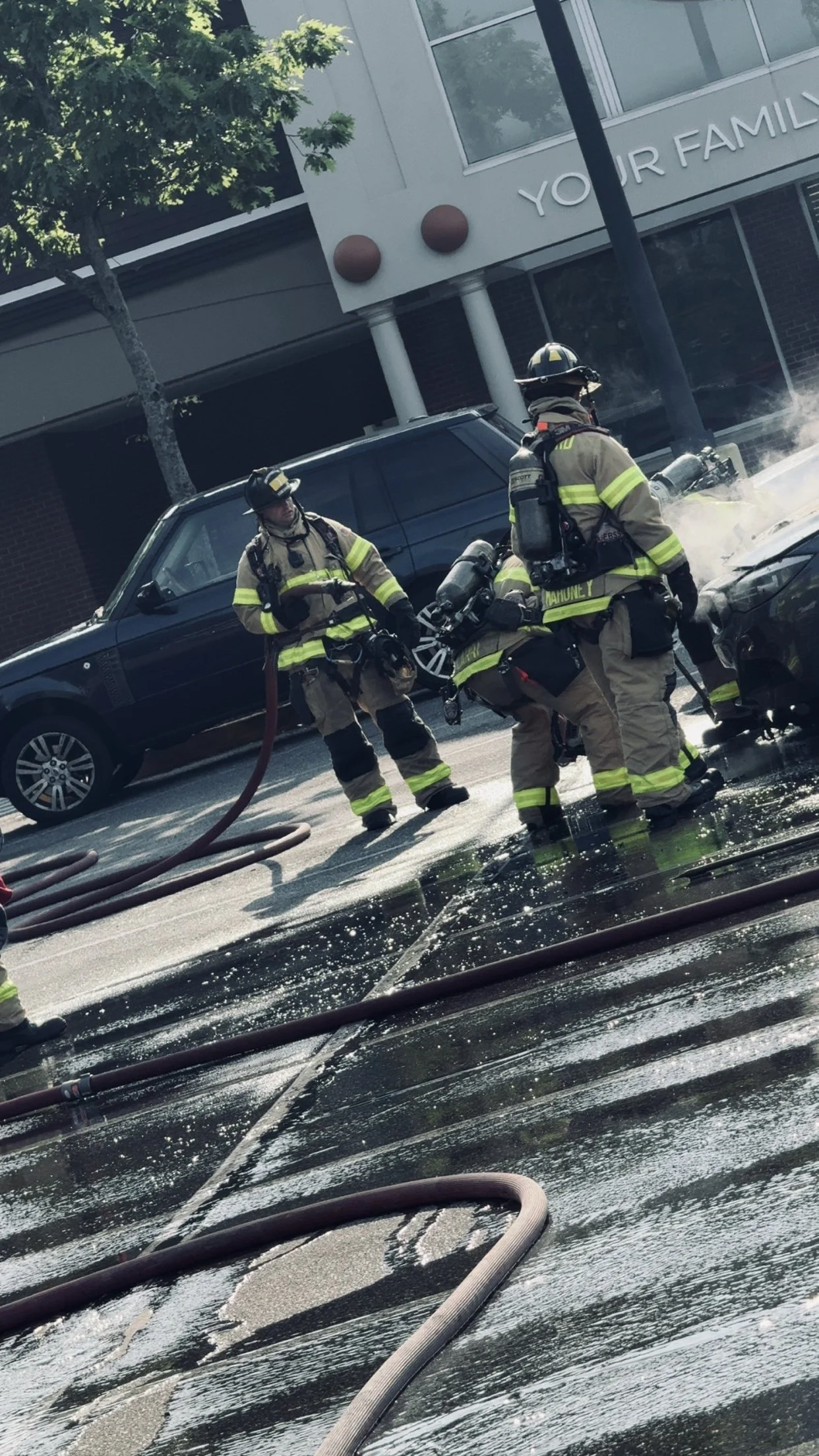 Firefighters in gear working at the scene of a car fire, with hoses on the ground and water spraying onto the vehicle.