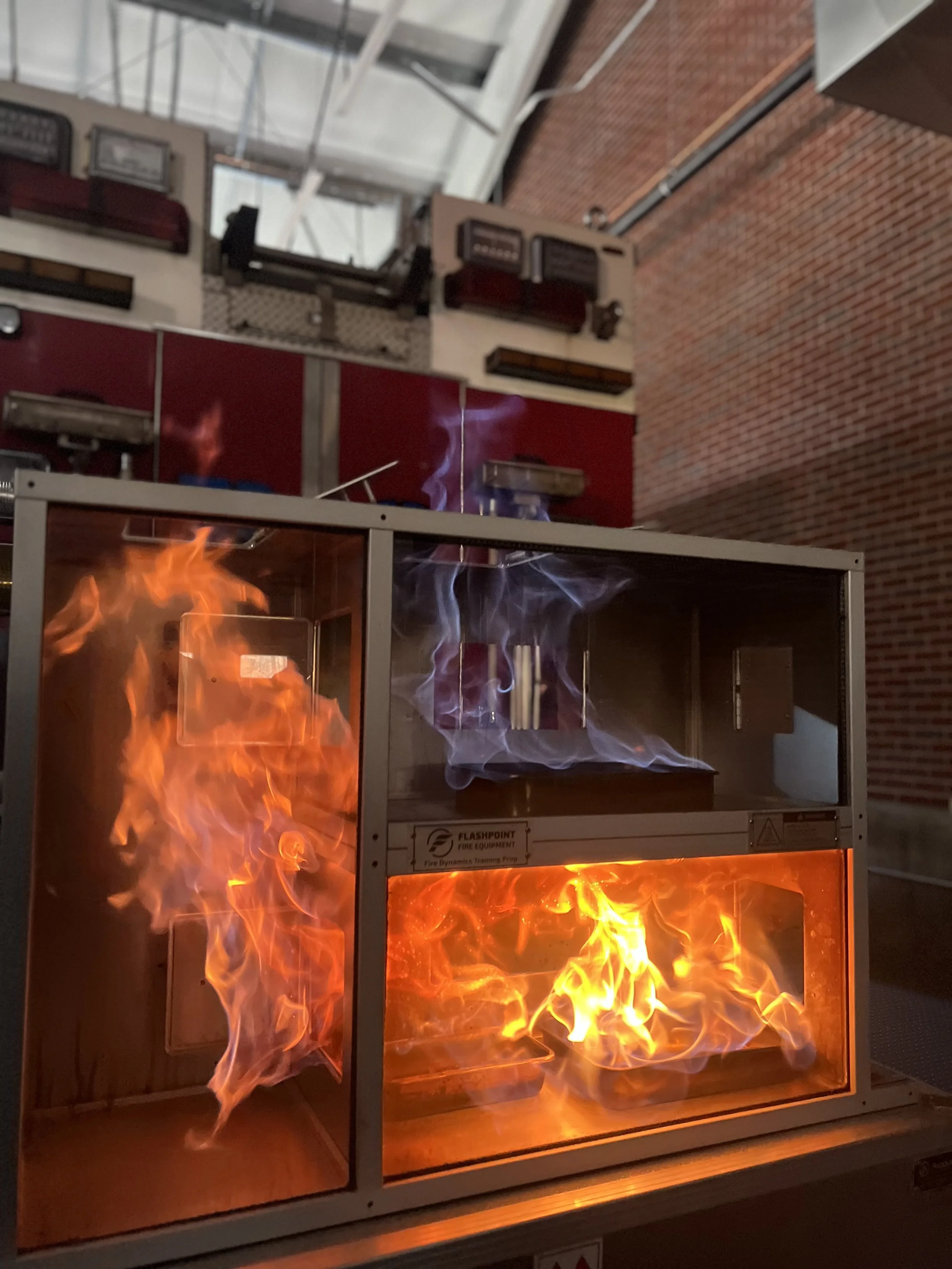A fire safety training device with visible flames inside a glass enclosure, organized in a room with brick and glass walls.