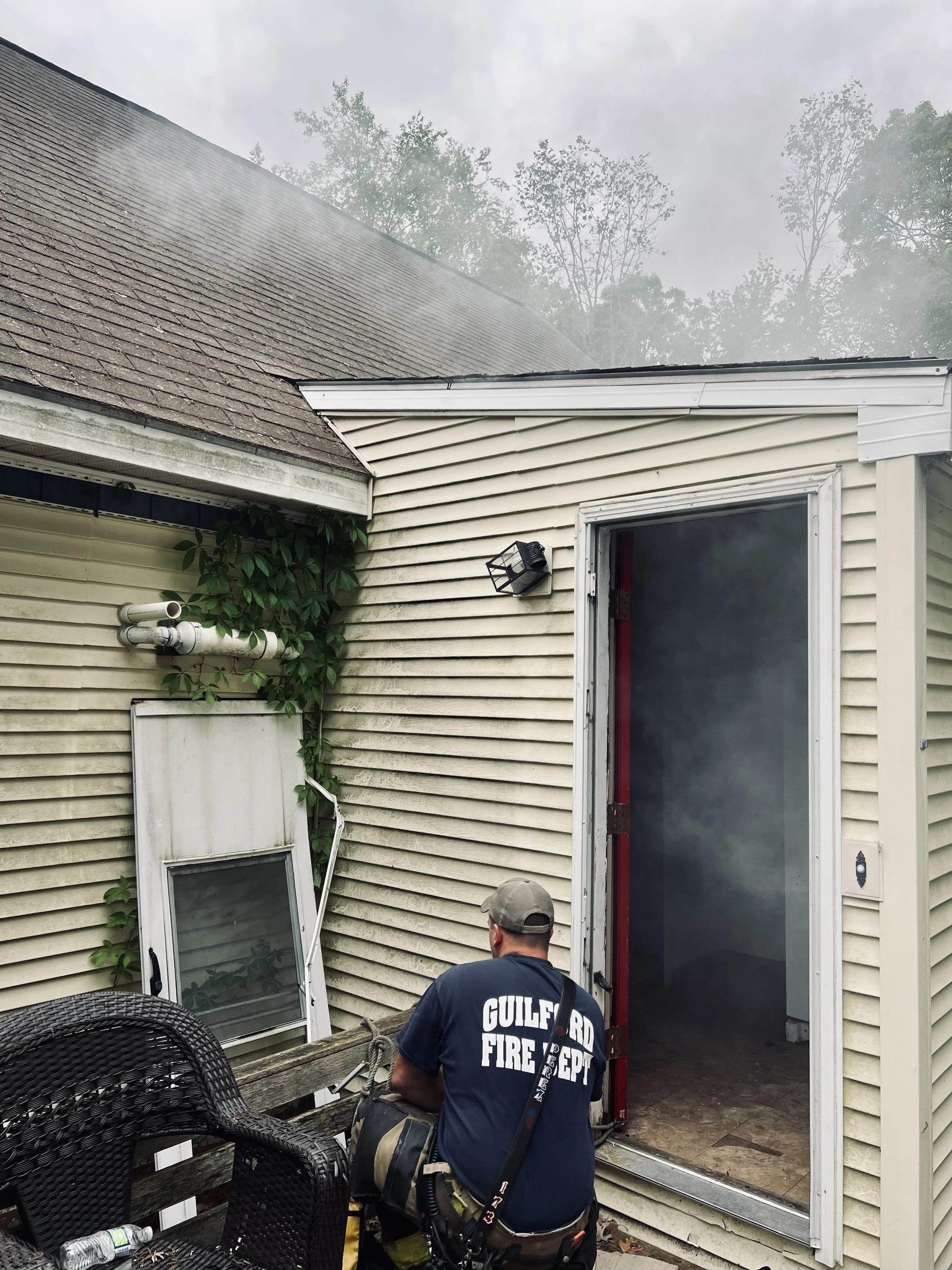 A firefighter from the Gulfport Fire Department stands near the back door of a house, which has slight damage to the door frame. There is a person with a backpack and equipment in the foreground, and outdoor furniture with a water bottle on it. The house has beige siding and a vent on the wall, with some green vines growing nearby. The roof shows signs of smoke or fire damage.