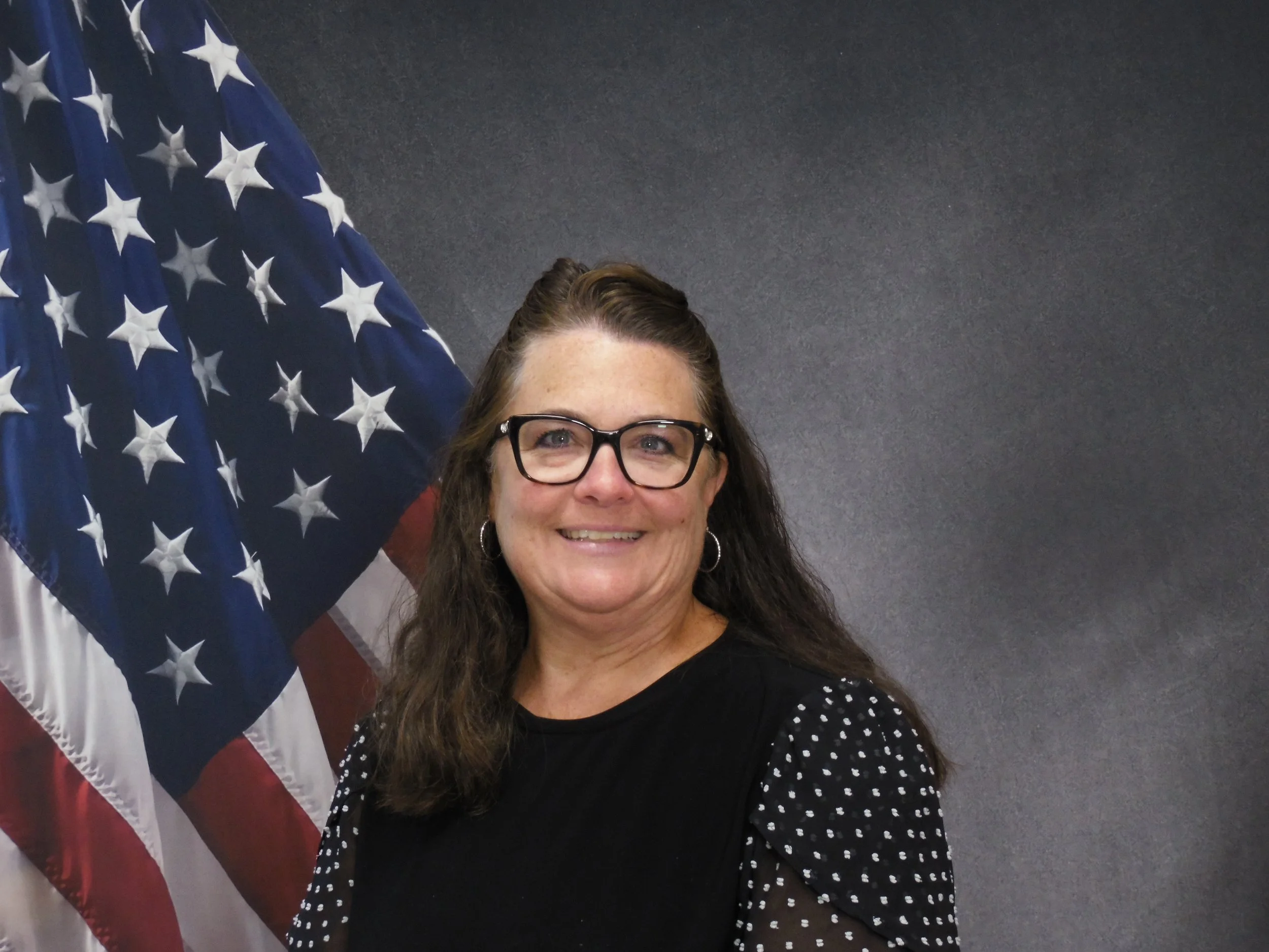 Smiling woman with glasses standing in front of an American flag and a dark wall background.