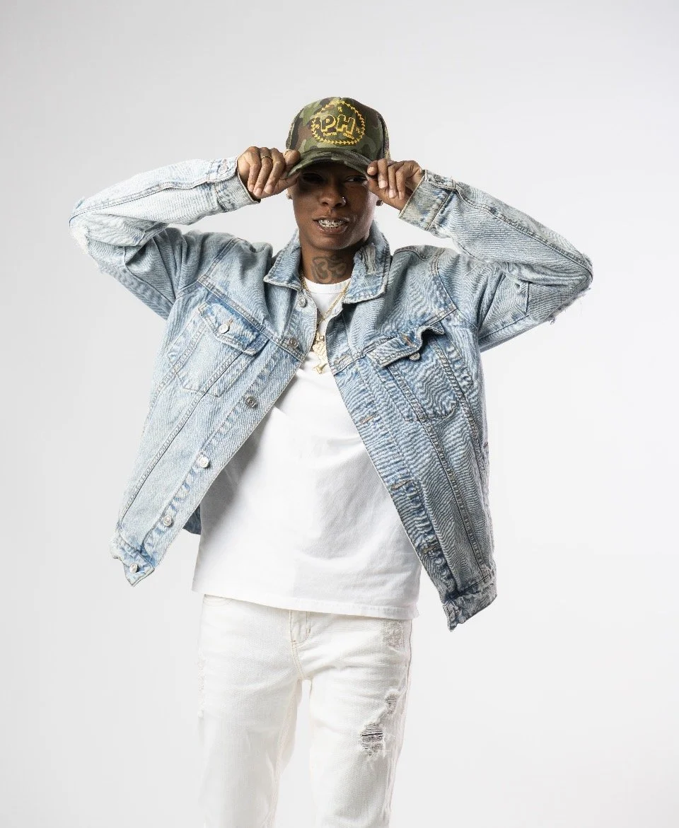 Young man wearing a denim jacket, white t-shirt, and white ripped jeans, holding a camouflage cap on his head, standing against a plain white background.