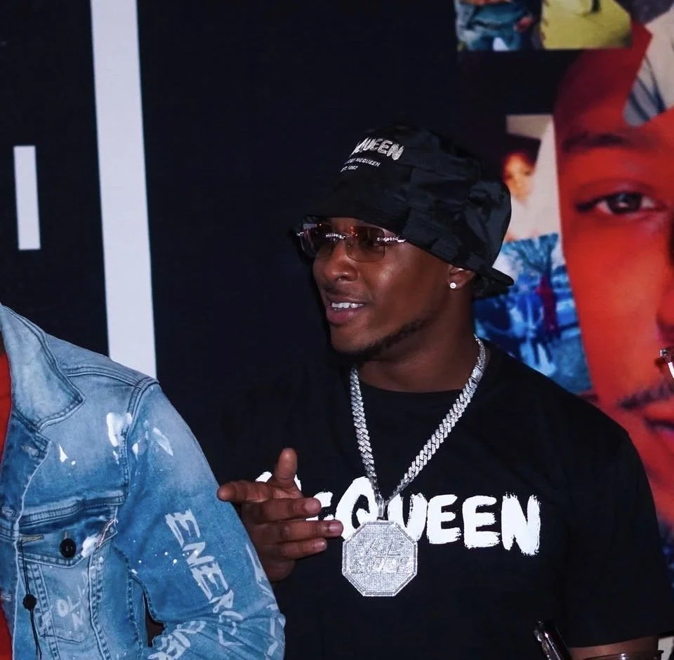A young man wearing a black bucket hat, sunglasses, and a black T-shirt with the word QUEEN, posing with his hand making a finger gun gesture. He has a large diamond necklace and stud earrings. In the background, there is a colorful poster.