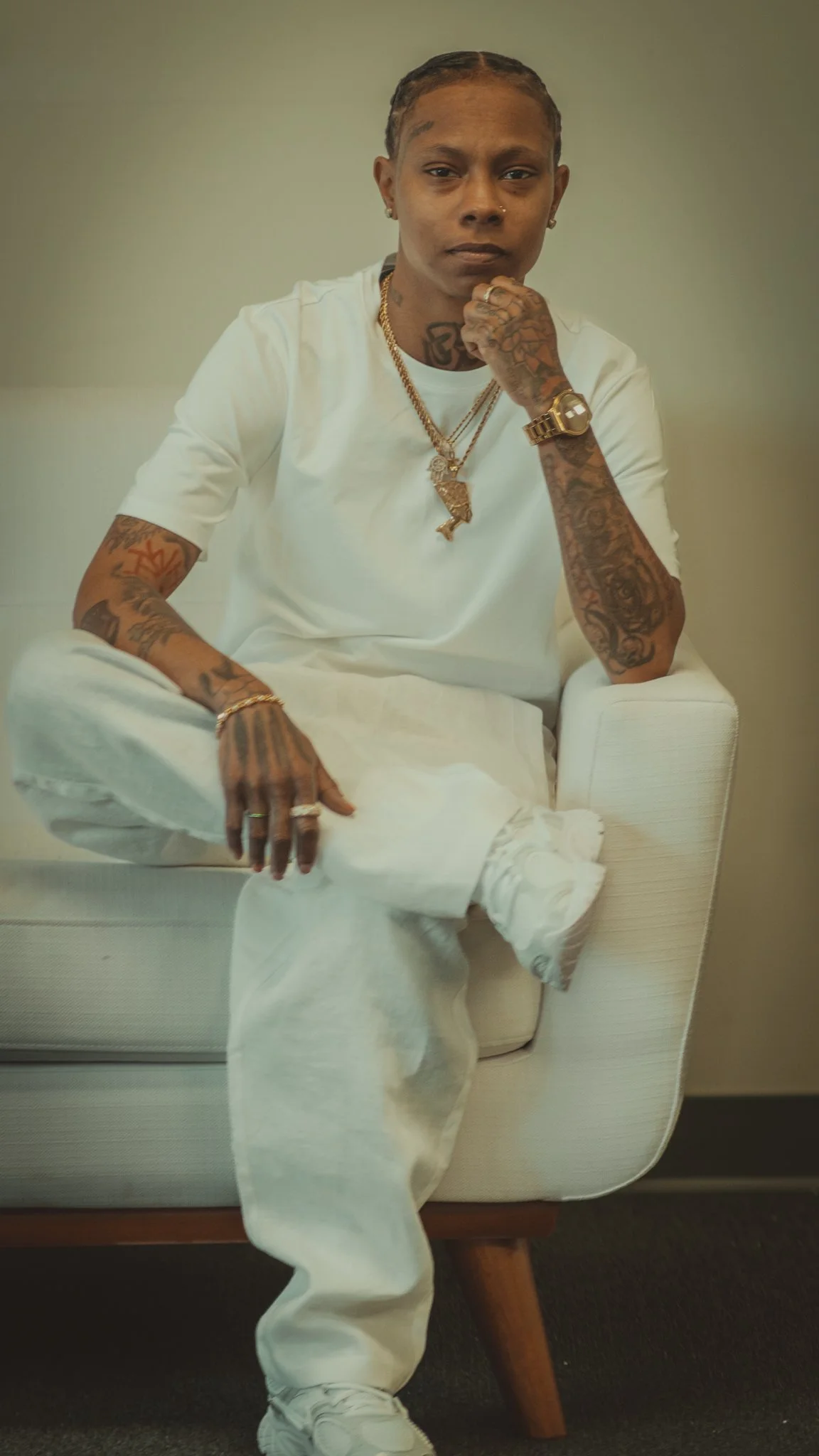 Young man wearing a denim jacket, white t-shirt, and white ripped jeans, holding a camouflage cap on his head, standing against a plain white background.