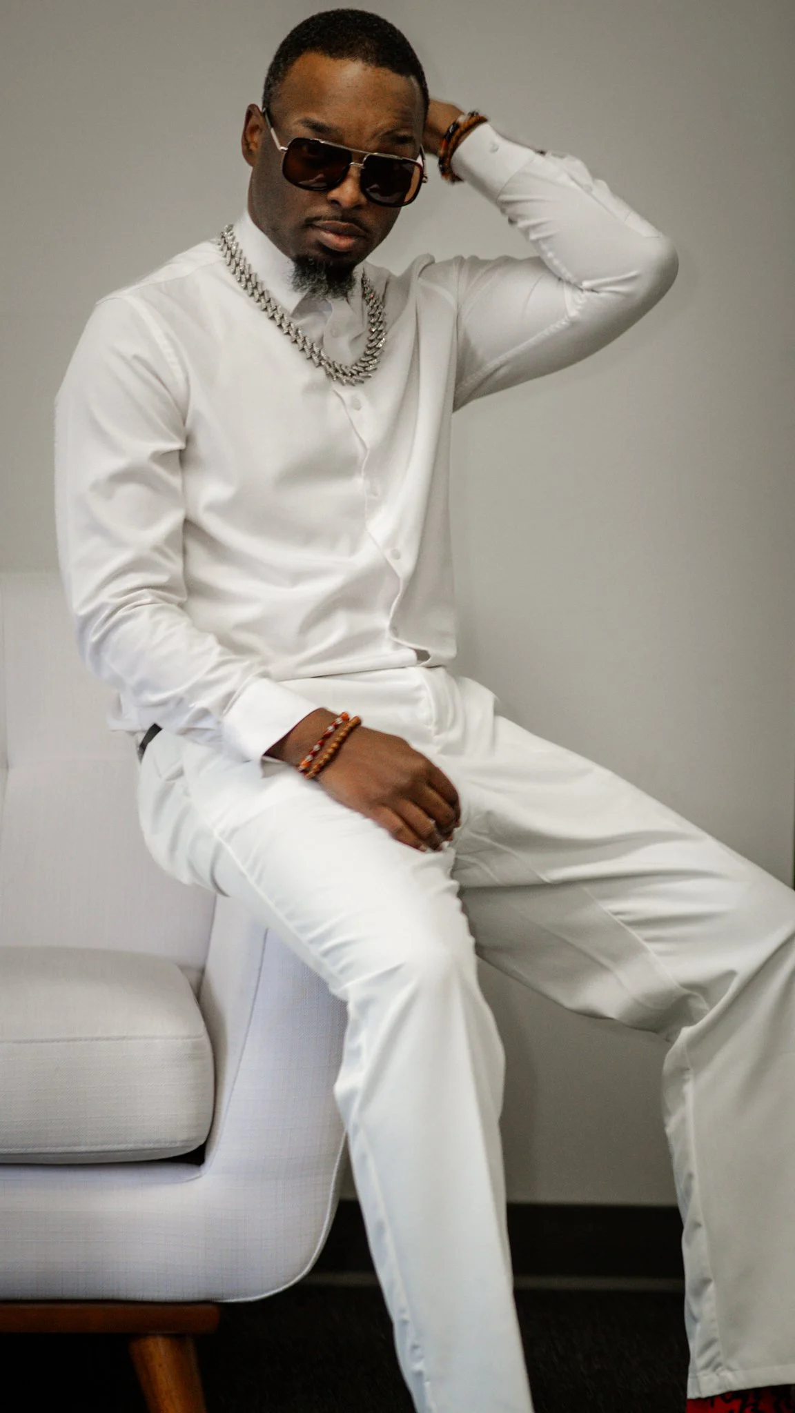 A man wearing a black hat, reflective sunglasses, a black sweater, and jewelry, sitting at a table with a bouquet of red roses and illuminated mirrors in the background.