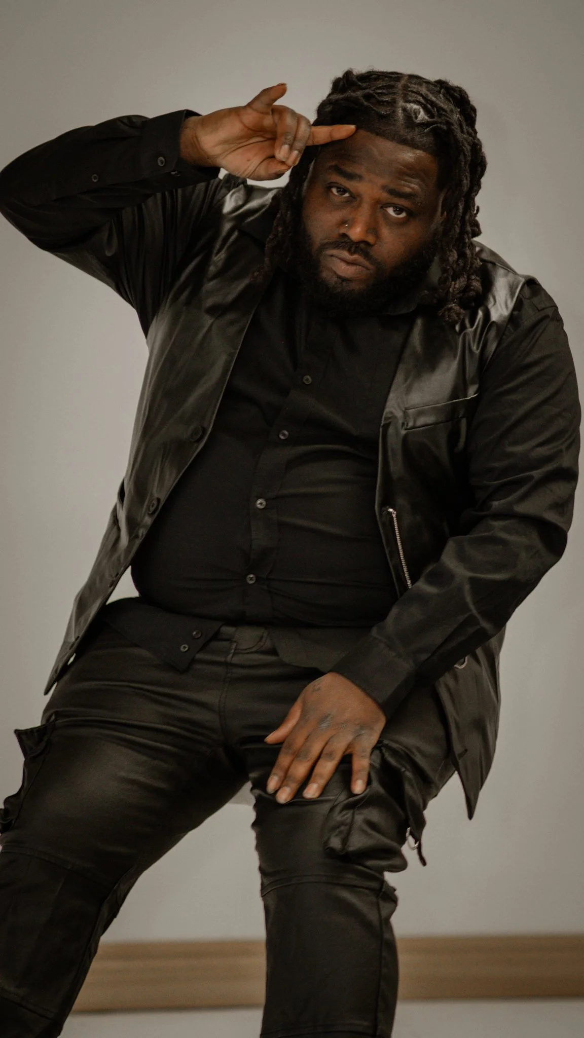 A man with styled dreadlocks and a beard, wearing sunglasses and a blue long sleeve shirt with a black vest, sitting at a table with his arms crossed.