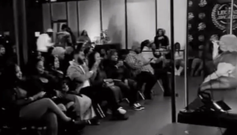 People seated and standing in a room, appearing to be attending a presentation or event, with a speaker at the front.