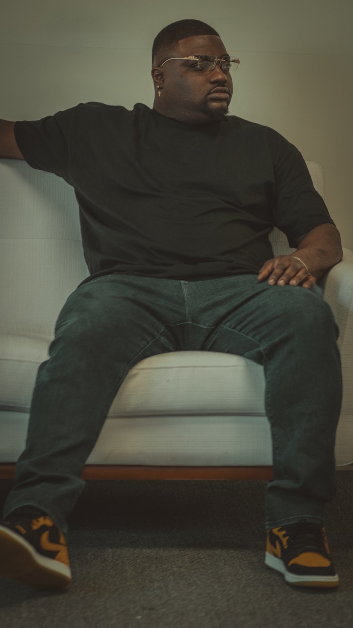 A man wearing dark sunglasses, a graphic t-shirt with the word 'COOL' in blue, yellow cargo pants, and white sneakers, standing outside at night with purple and black background lighting.