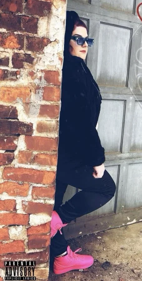 Woman with dark hair and sunglasses leaning against a wall, wearing a black jacket, black pants, and bright pink shoes