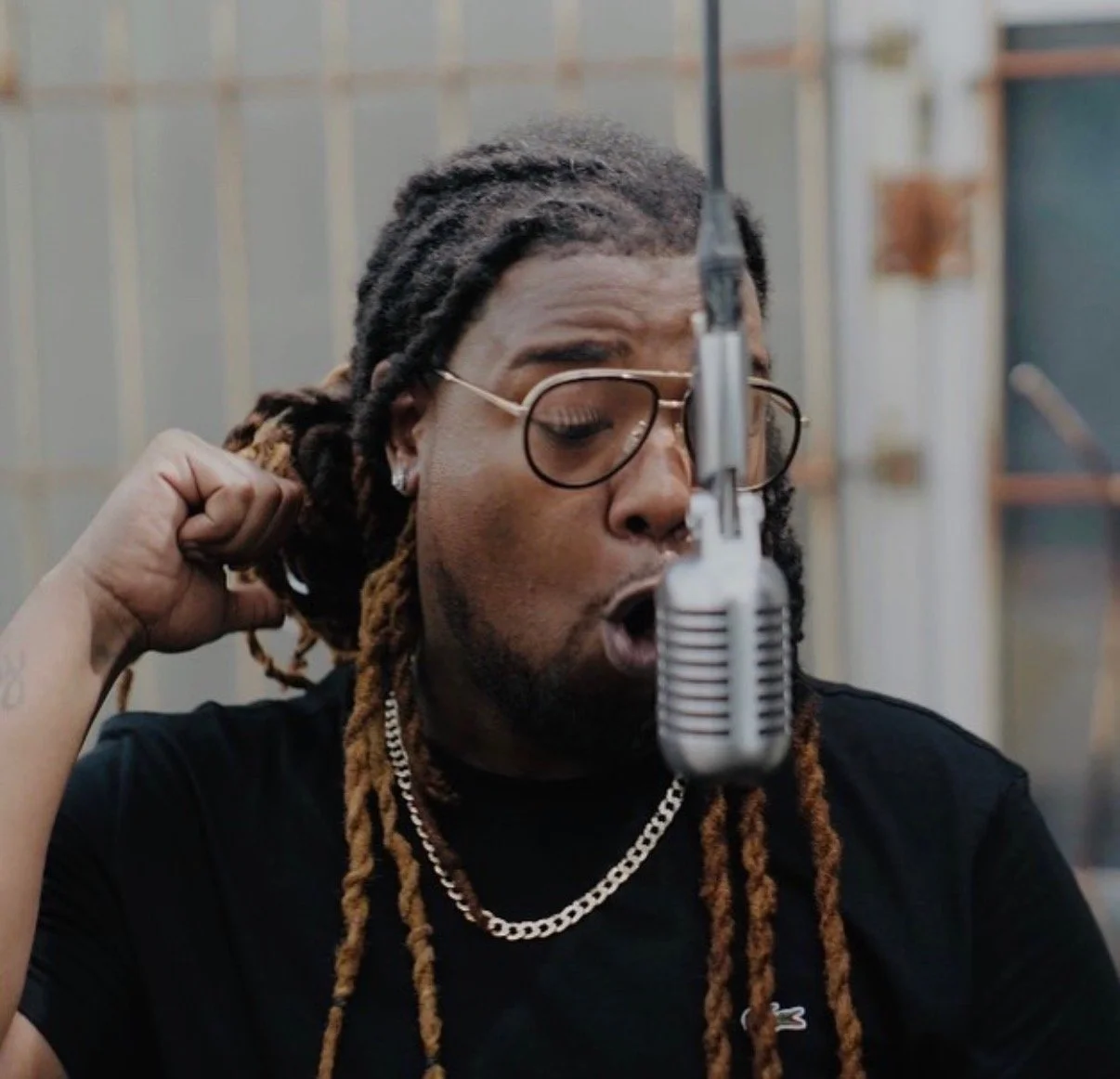 A man with long dreadlocks and glasses singing into a microphone in a recording studio.