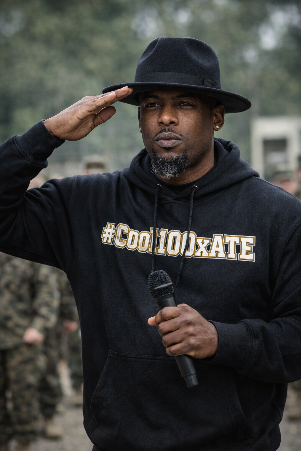 A man in a black hoodie with the hashtag '#COOL10XATE' and a black hat, saluting while holding a microphone outdoors.