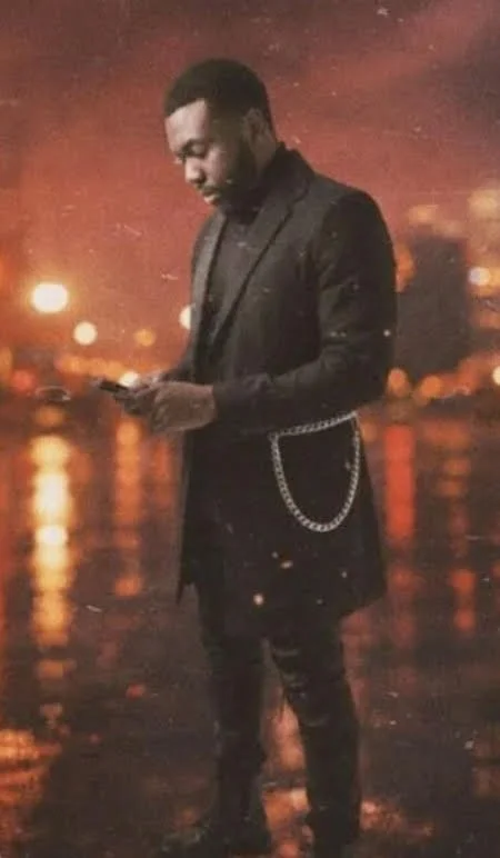 A man in a black suit with a chain pocket accessory is standing outdoors at night, looking at his phone, with blurred city lights and reflections on wet ground in the background.