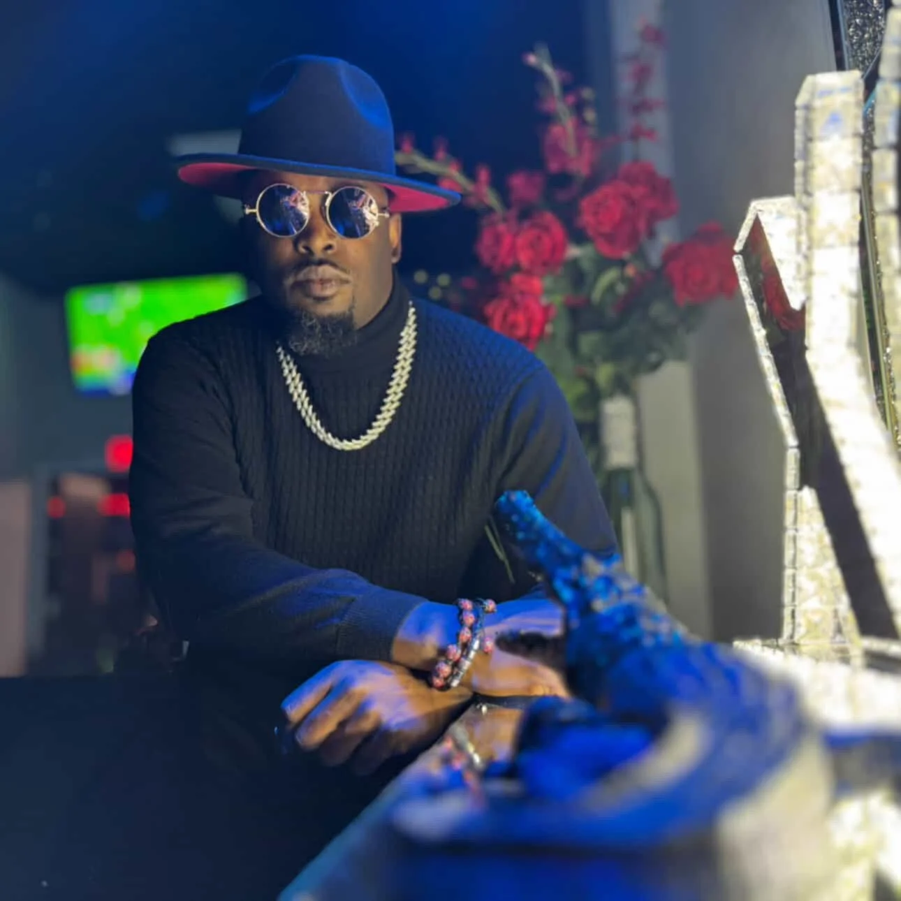A man wearing a black hat, reflective sunglasses, a black sweater, and jewelry, sitting at a table with a bouquet of red roses and illuminated mirrors in the background.