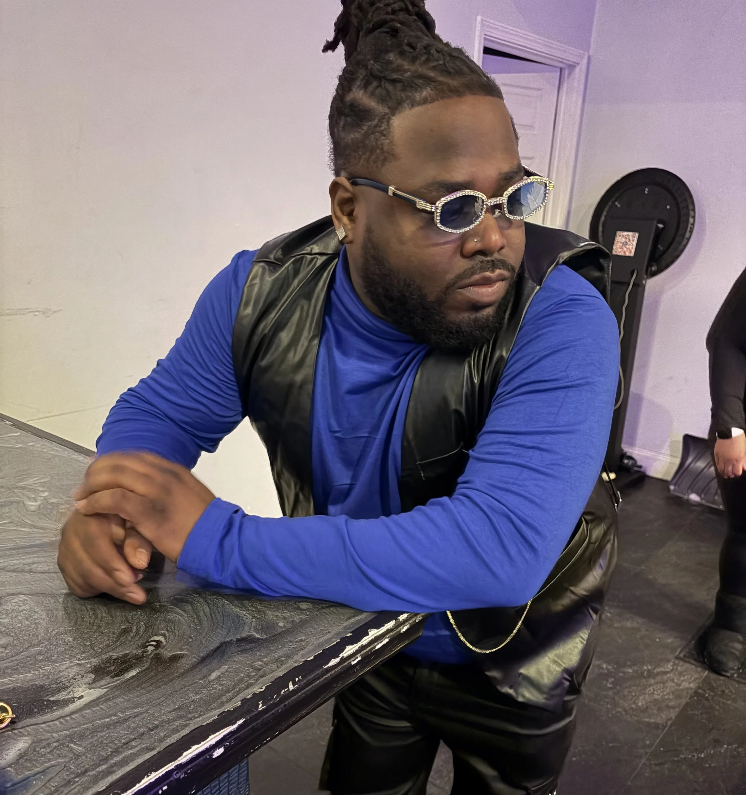 A man with styled dreadlocks and a beard, wearing sunglasses and a blue long sleeve shirt with a black vest, sitting at a table with his arms crossed.