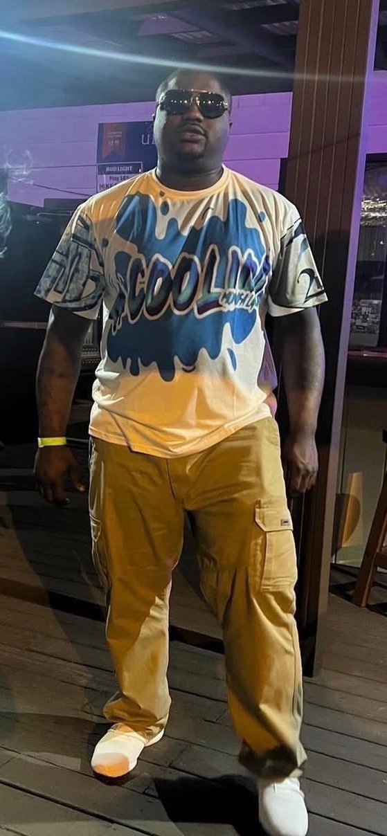 A man wearing dark sunglasses, a graphic t-shirt with the word 'COOL' in blue, yellow cargo pants, and white sneakers, standing outside at night with purple and black background lighting.