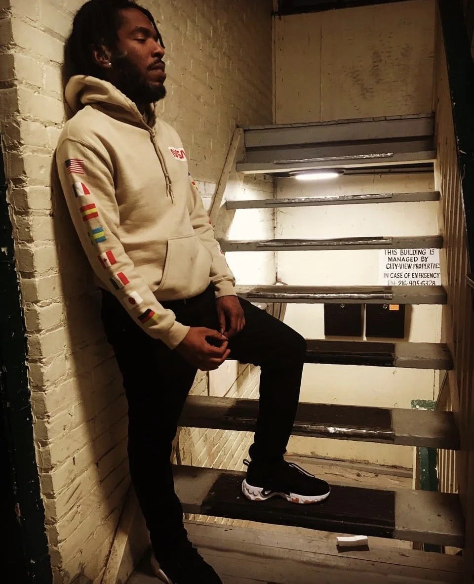 A young man with dreadlocks and a beard, dressed in a beige hoodie with flags and an 'NASA' logo, leaning against a brick wall near stairs inside a building.