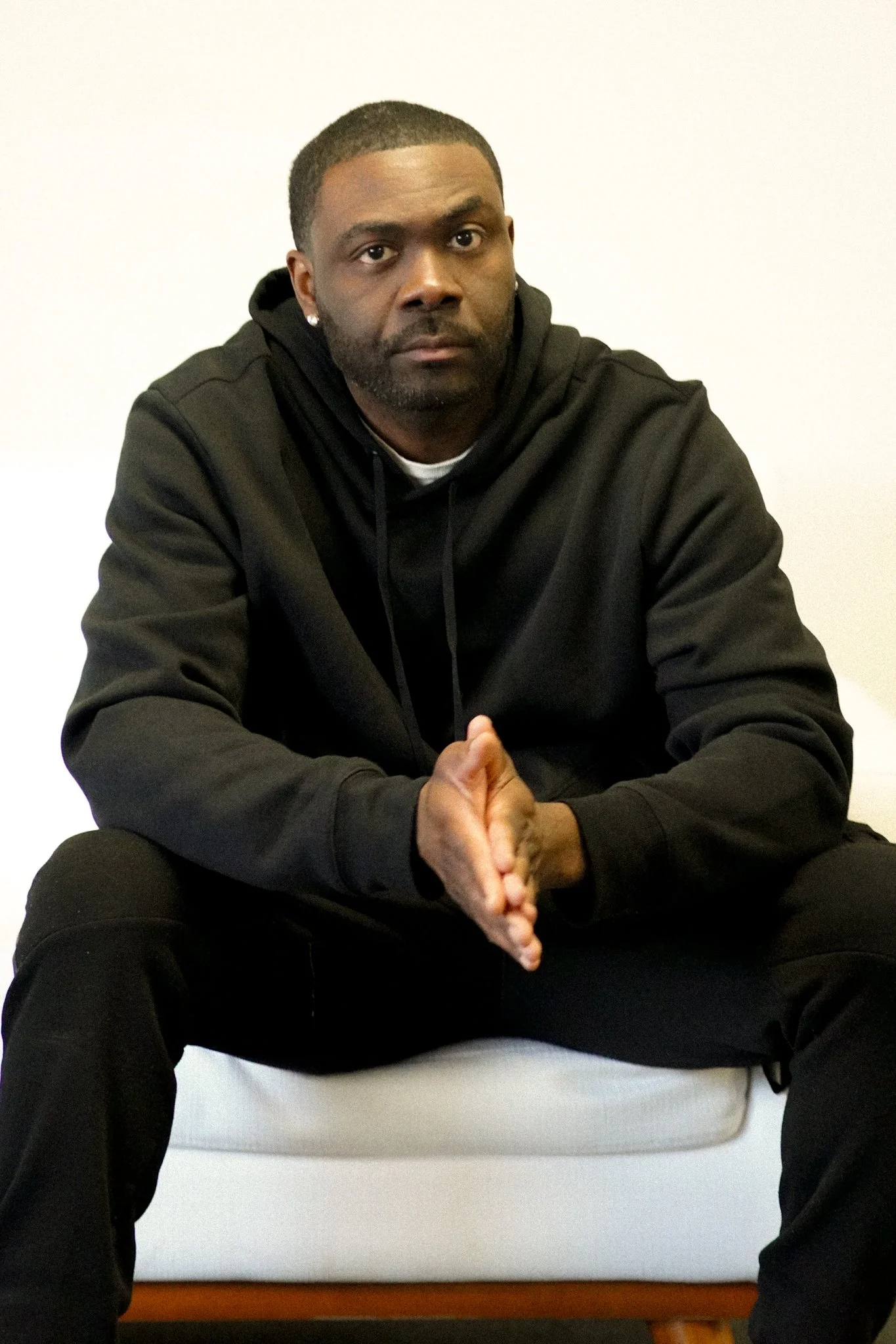 A man in a black suit with a chain pocket accessory is standing outdoors at night, looking at his phone, with blurred city lights and reflections on wet ground in the background.
