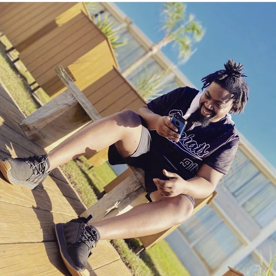 A man with dreadlocks smiling and looking at his phone while sitting on a wooden bridge outdoors. There are wooden fences and palm trees in the background on a sunny day.