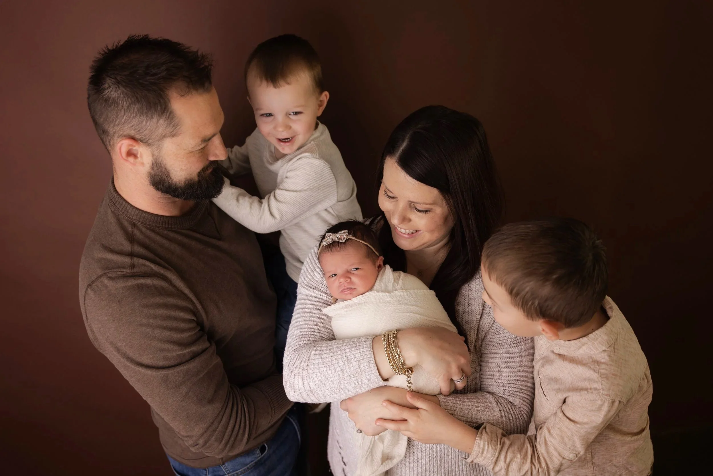 A family of six people, including one man, one woman, and three children, is gathered together, smiling and interacting with each other against a dark background.