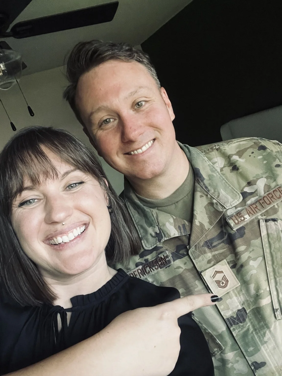 A smiling woman with dark hair and a piercing taking a selfie with a man in military uniform, pointing to his uniform badge.