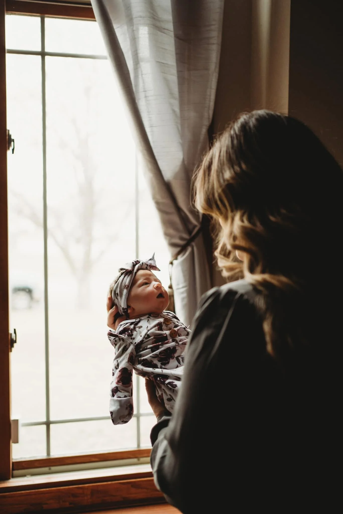 A woman with long hair holding a baby girl by a large window. The baby, wearing a floral jacket and headband, looks up at the woman. The window has white curtains, and outside is a blurred view of trees and sky.