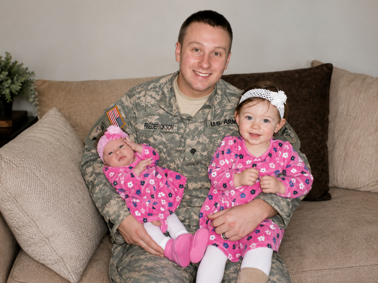 A soldier in uniform holding two young girls, one older and one younger, on a beige couch in a living room. The girls are wearing matching pink floral pajamas. The oldest girl has a white headband, and the younger girl has a pink headband with a flower.