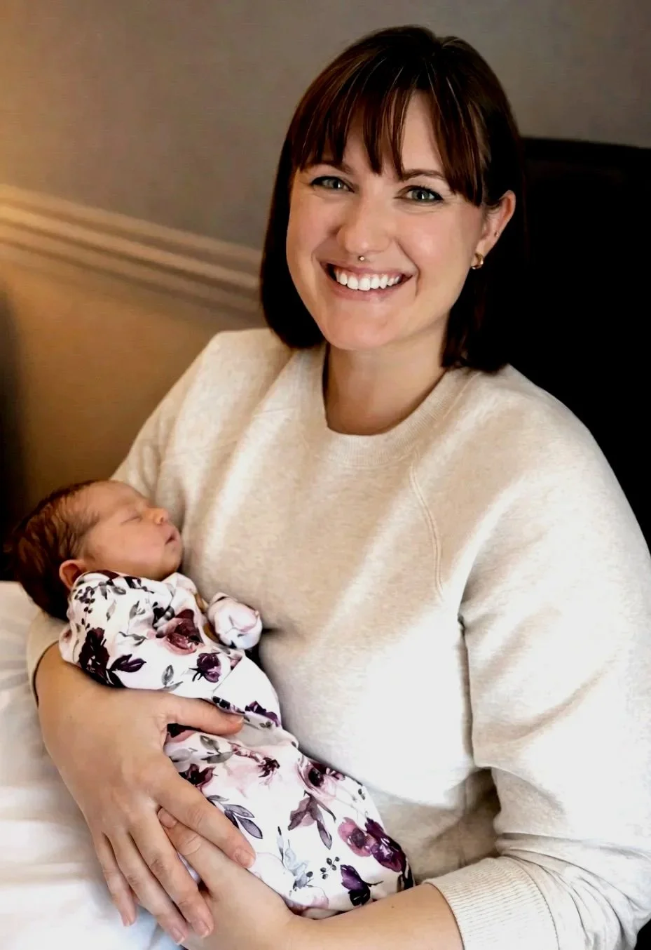 A woman with short brown hair holding a sleeping newborn baby wrapped in a floral blanket.