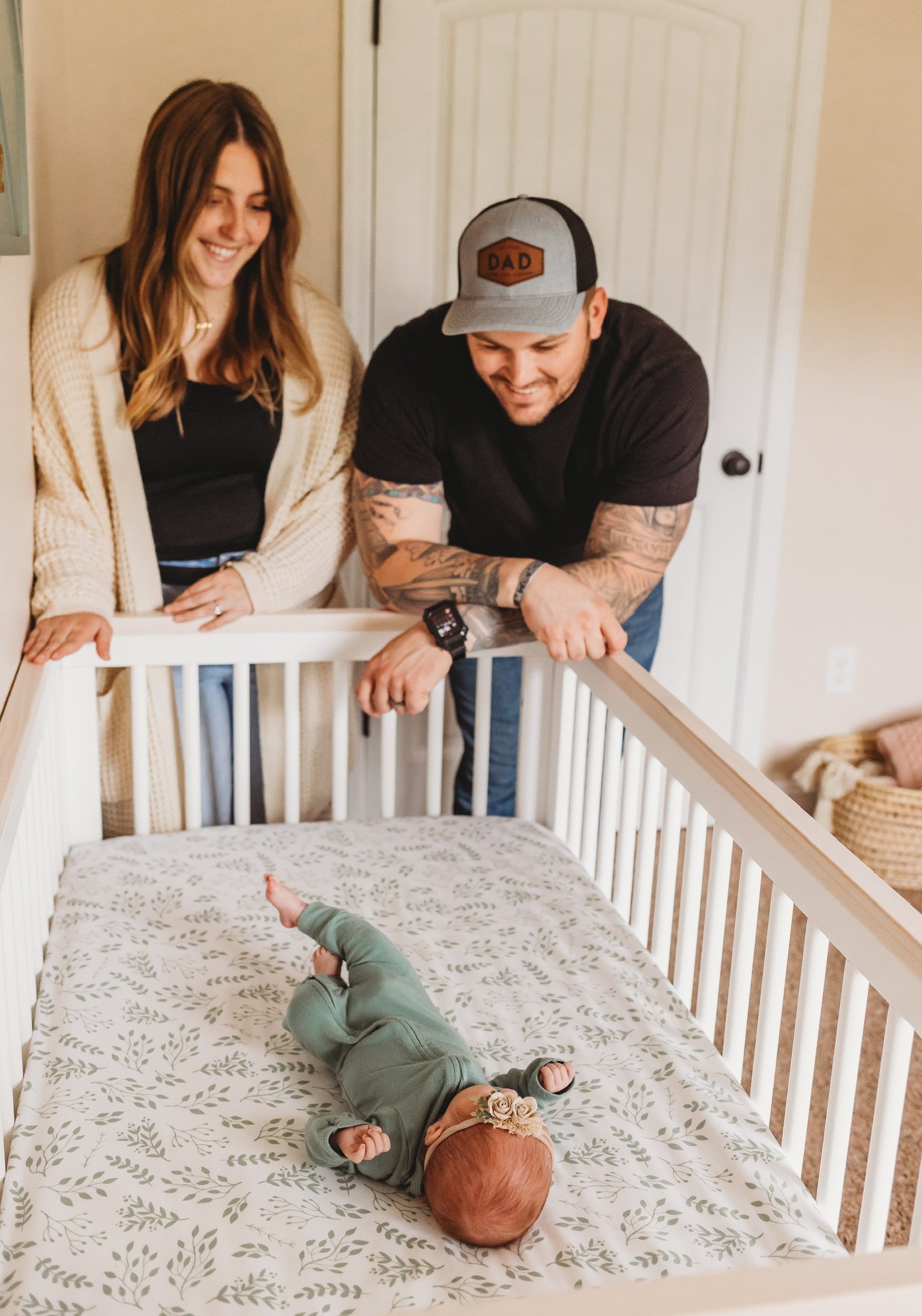 A woman and a man are looking at a baby lying in a crib. The woman is smiling and wearing a beige cardigan, while the man with tattoos on his arms is leaning on the crib, smiling and wearing a cap that says "DAD." The baby is lying on its back in a crib with a patterned sheet, wearing greenish-brown clothes and a headband with a flower decoration.