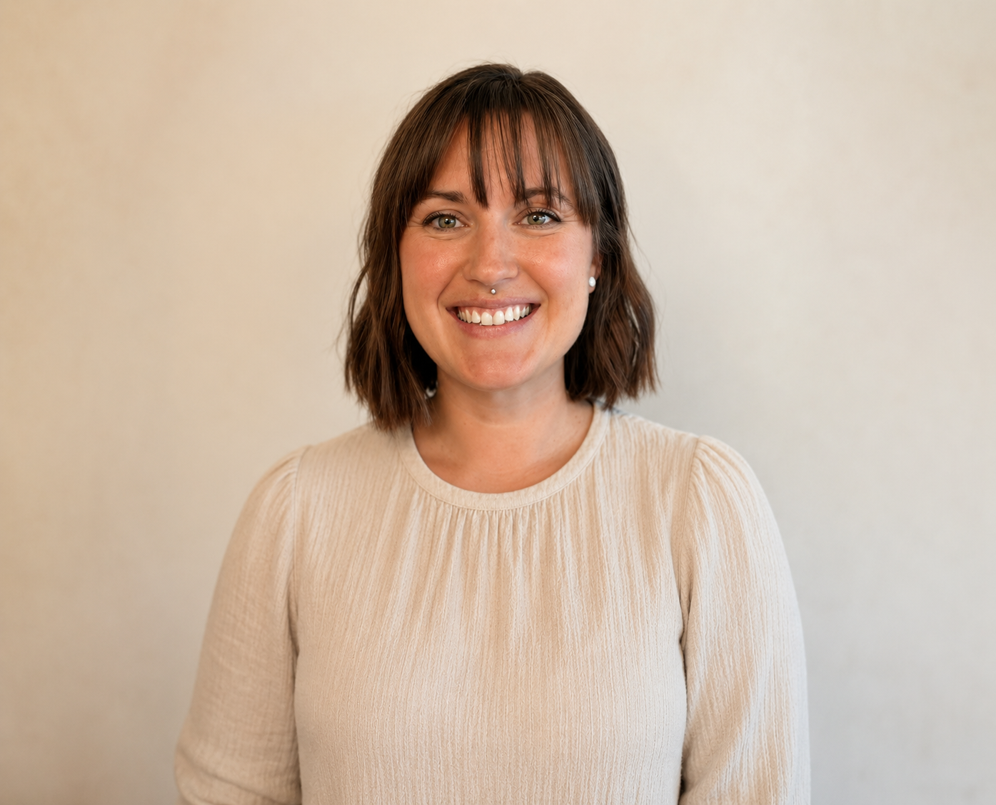 A woman with shoulder-length brown hair and bangs, wearing a beige sweater, smiling at the camera against a plain light-colored background.