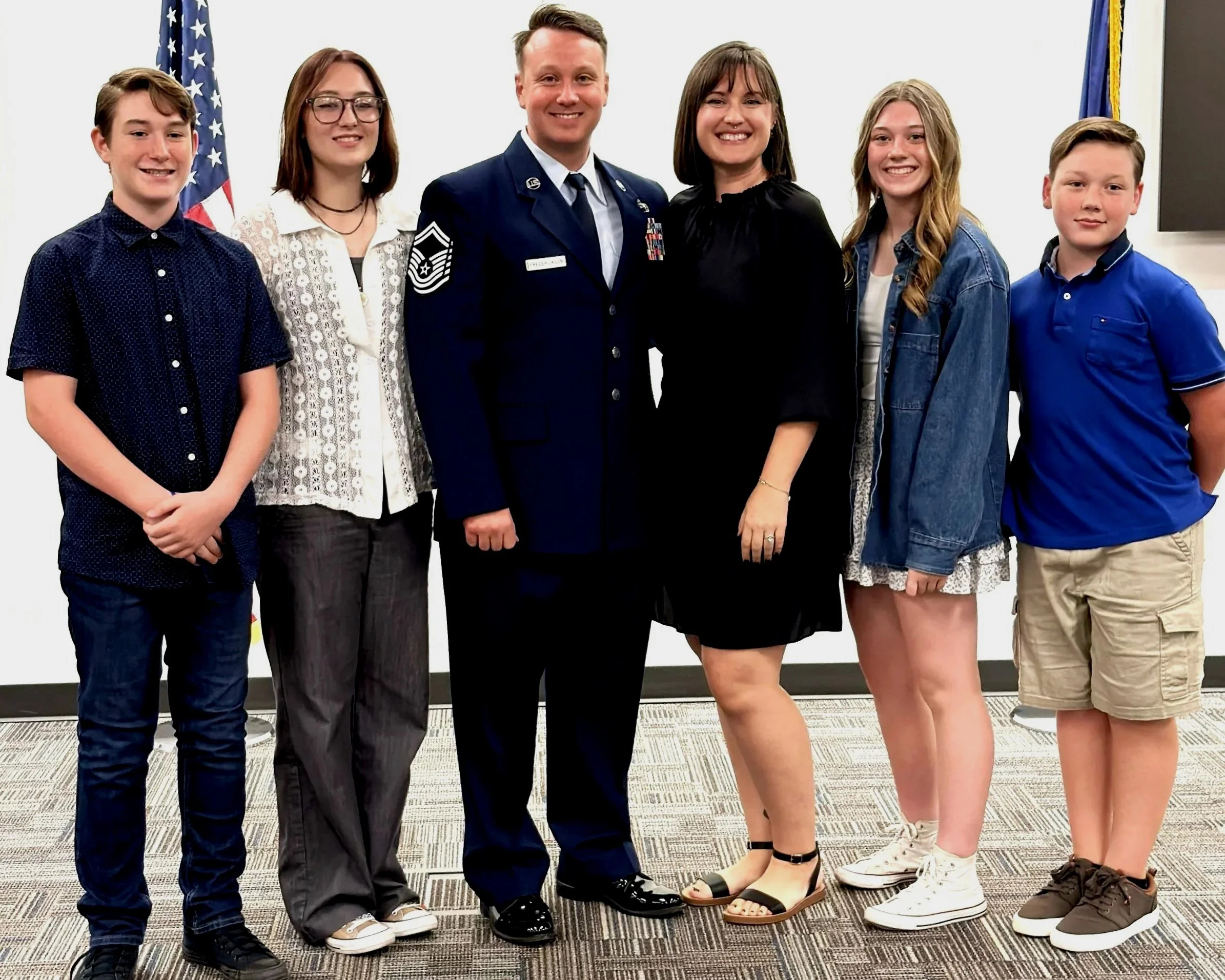 Group of six young people standing together indoors, with two American flags in the background. One person is in a military uniform, and the others are in casual clothing, smiling for the camera.