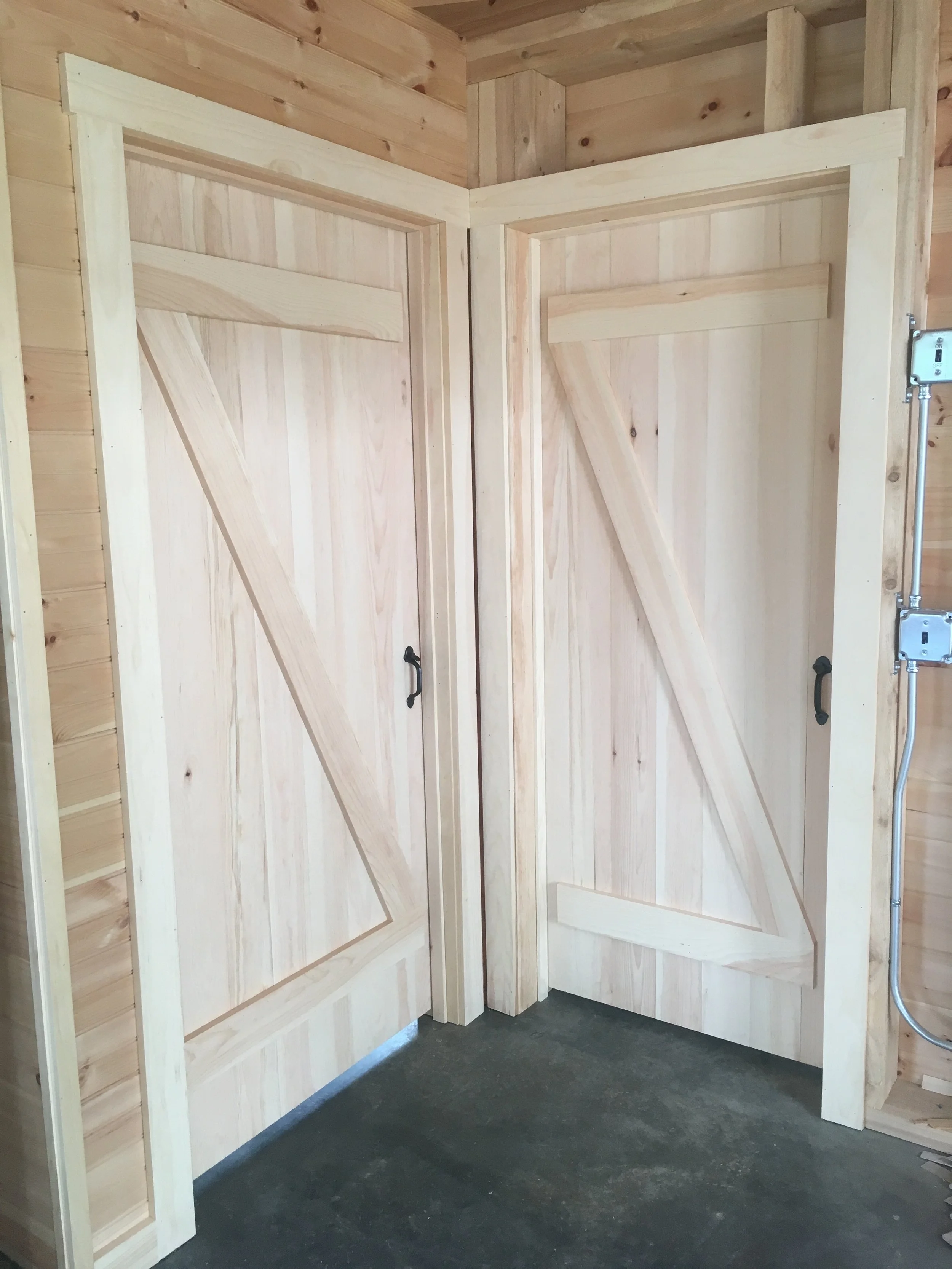 Corner of a room with unfinished wooden walls, two vertical wooden doors with diagonal planks and black handles, and exposed electrical wiring.