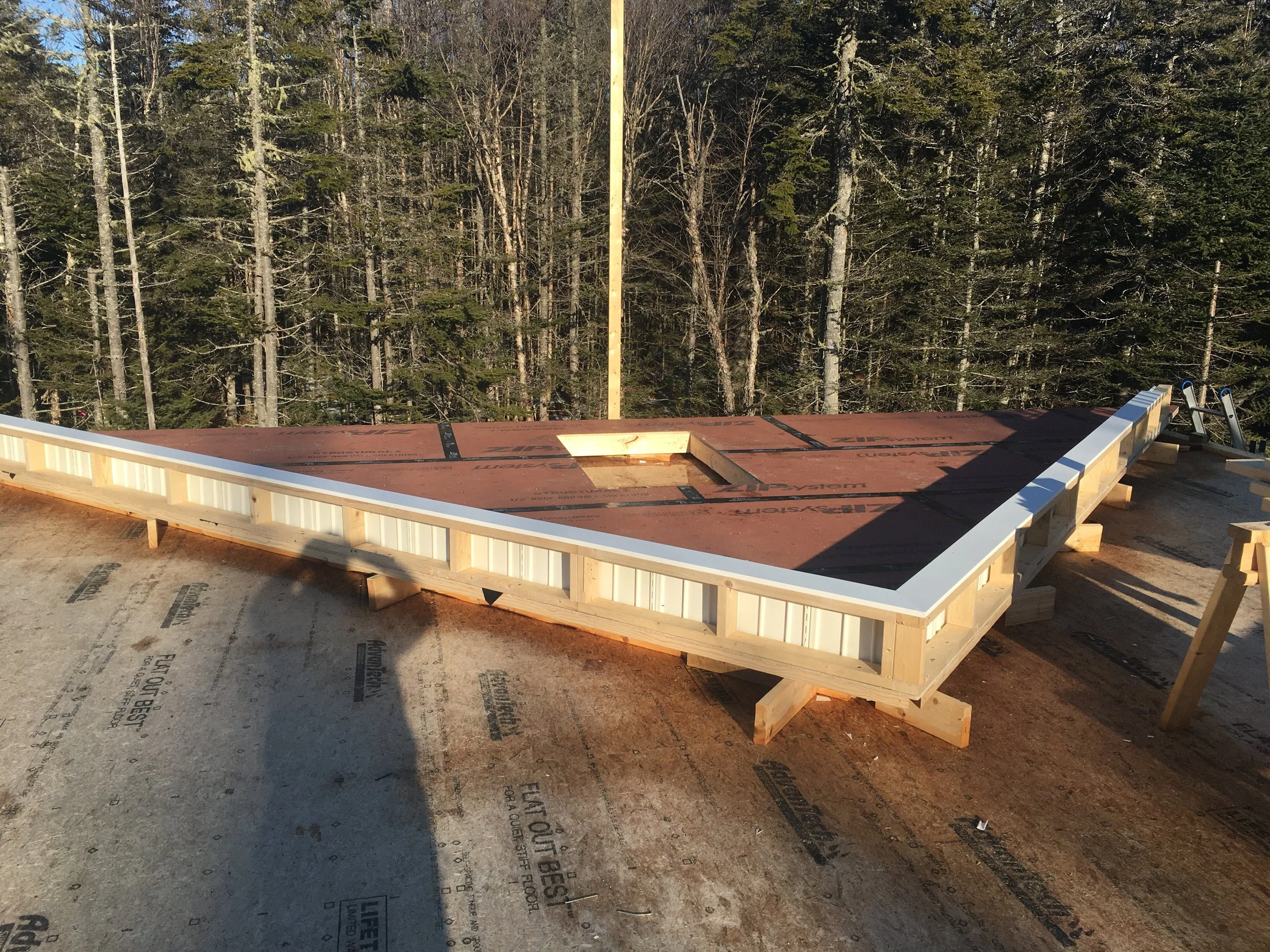 Construction site with a wooden framed platform, surrounded by a forest of tall trees in the background, sunlight casting shadows on the unfinished structure.