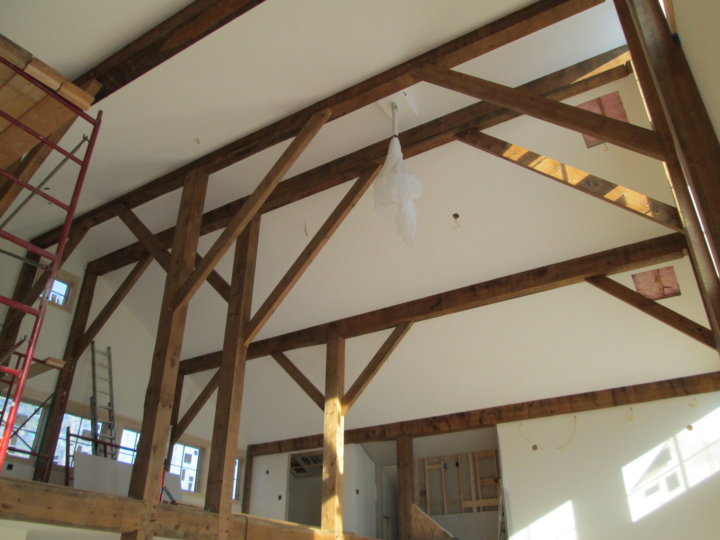 Interior of a house under construction, showing exposed wooden beams on the ceiling, windows, and construction scaffolding.