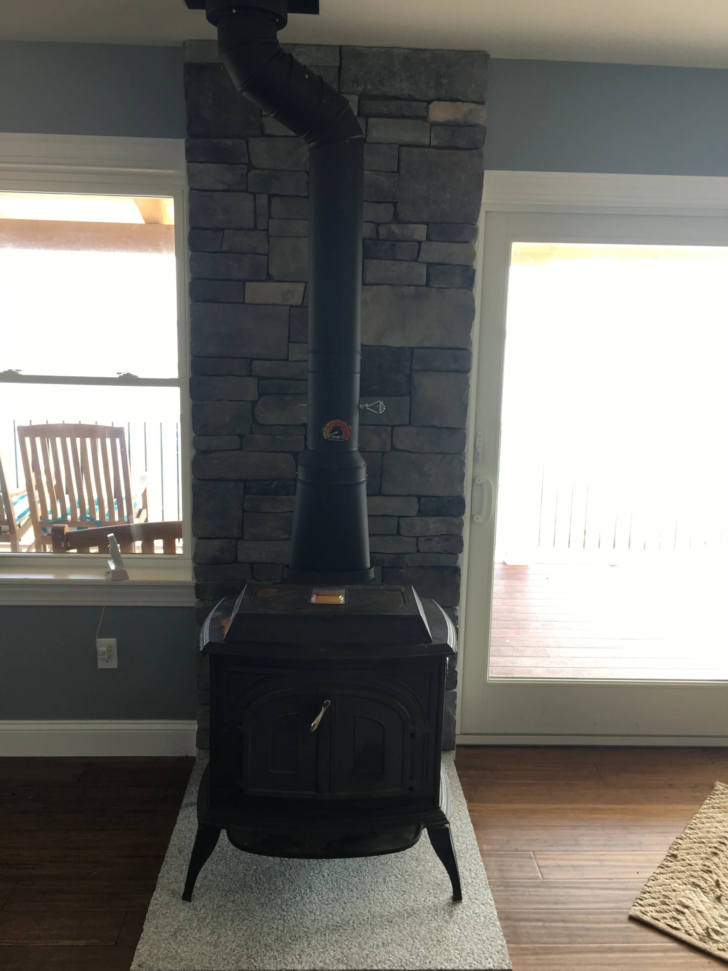 Black wood stove in front of a brick wall with a chimney pipe, positioned on a small rug in a room with hardwood floors, next to a window and a sliding glass door leading outside.