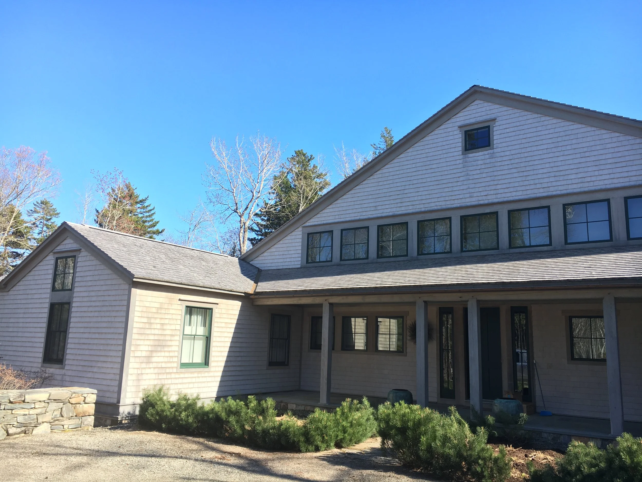 A house under construction with a beige exterior, multiple windows, and a sloped roof. The front porch is supported by wooden beams, with some construction materials visible. Trees and a clear blue sky are in the background.