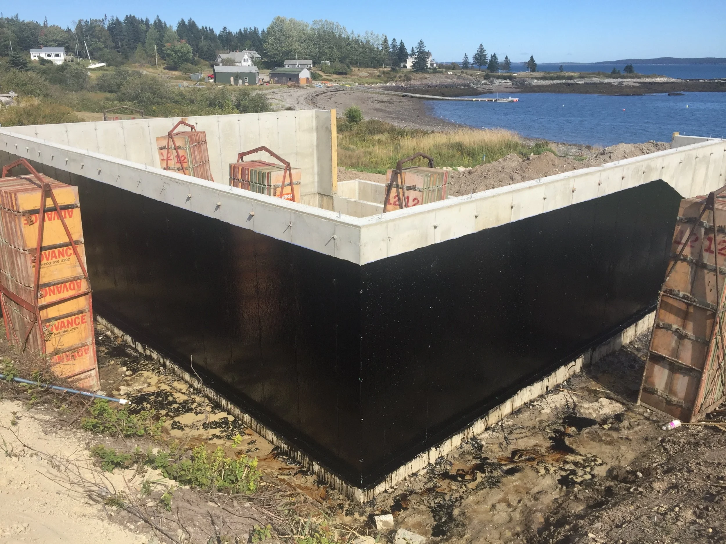 A construction site with concrete foundations and black waterproofing coating near a body of water, with trees and houses in the background.