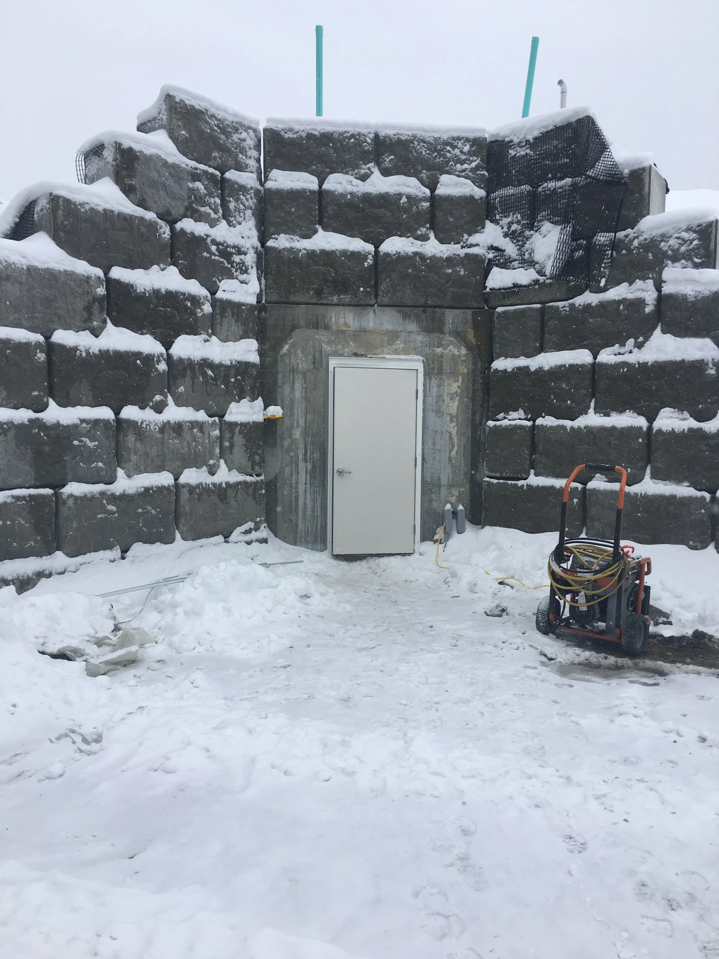 Snow-covered stone wall structure with a door in the center, situated outdoors in winter, with equipment and cables nearby.