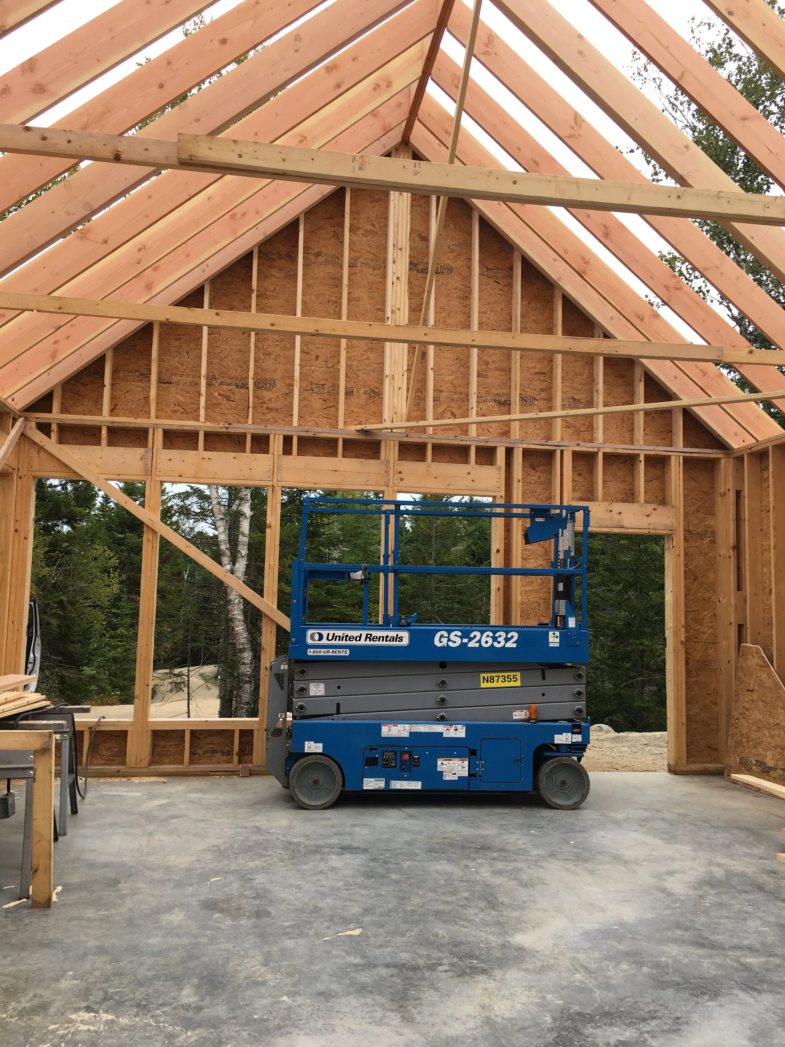 Interior of a house under construction with exposed wooden framing and roof trusses, and a blue scissor lift parked on a concrete floor.