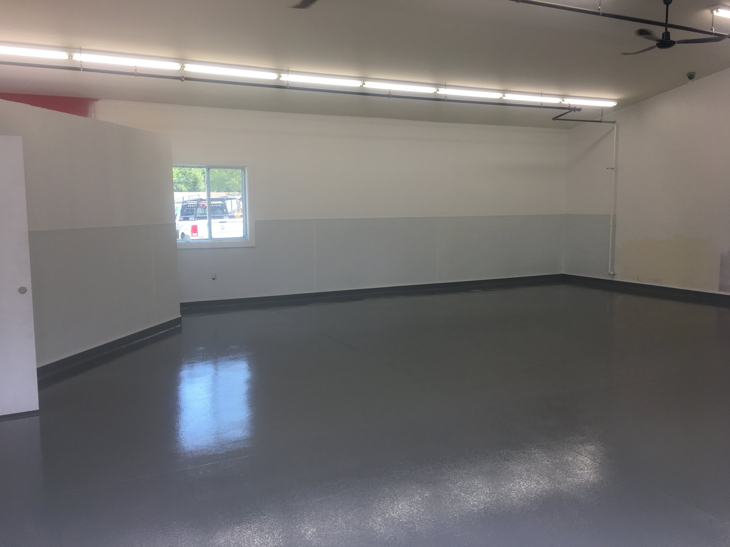 Empty room with gray floor, white walls, a window, fluorescent ceiling lights, and a ceiling fan.