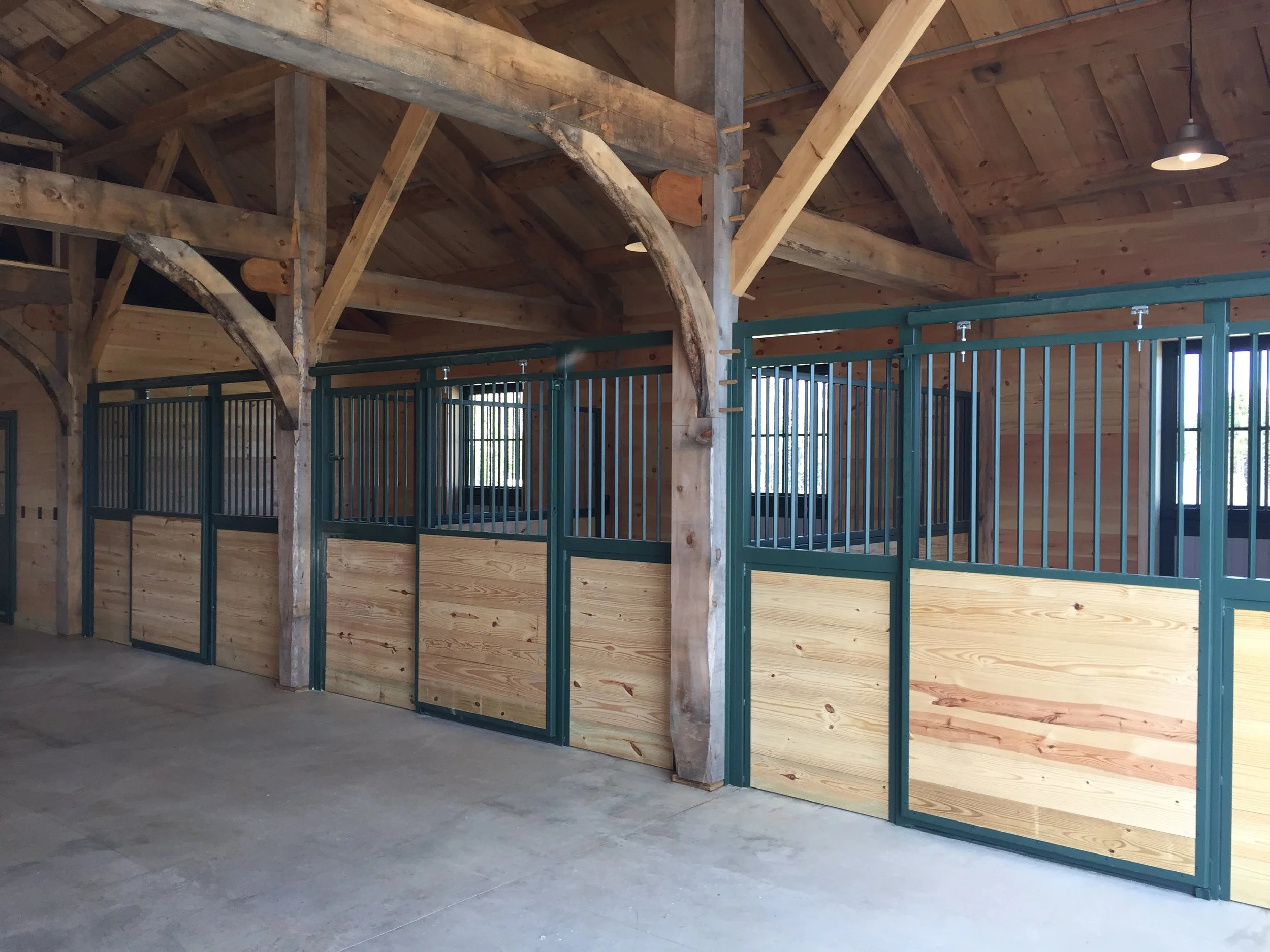 Interior of a barn showing a row of horse stalls with green metal frames and wooden panels, wooden beams, and windows.