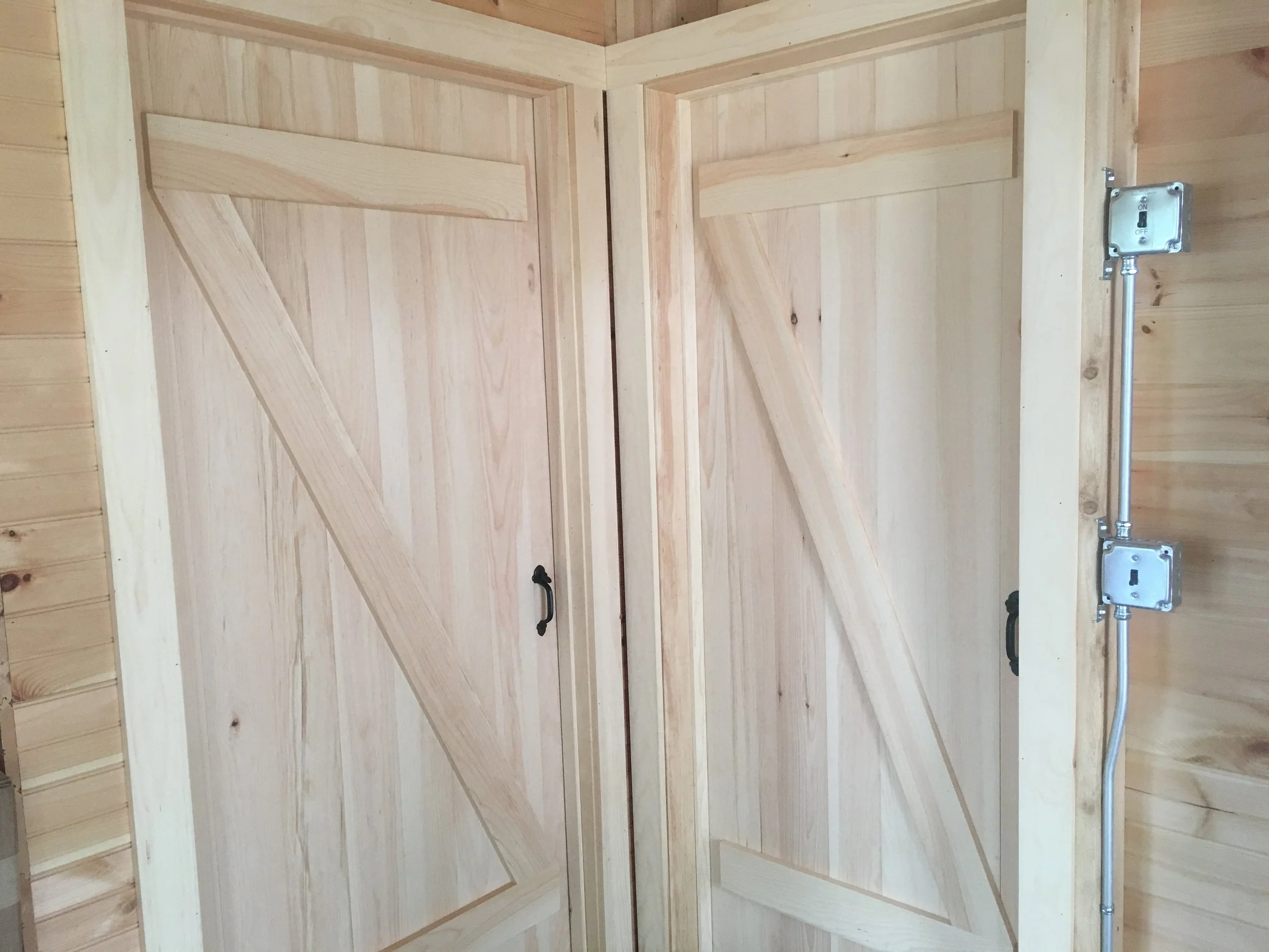 Two wooden barn-style doors with diagonal and horizontal panels, black handles, in a corner with wood-paneled walls and visible electrical conduit on the right.