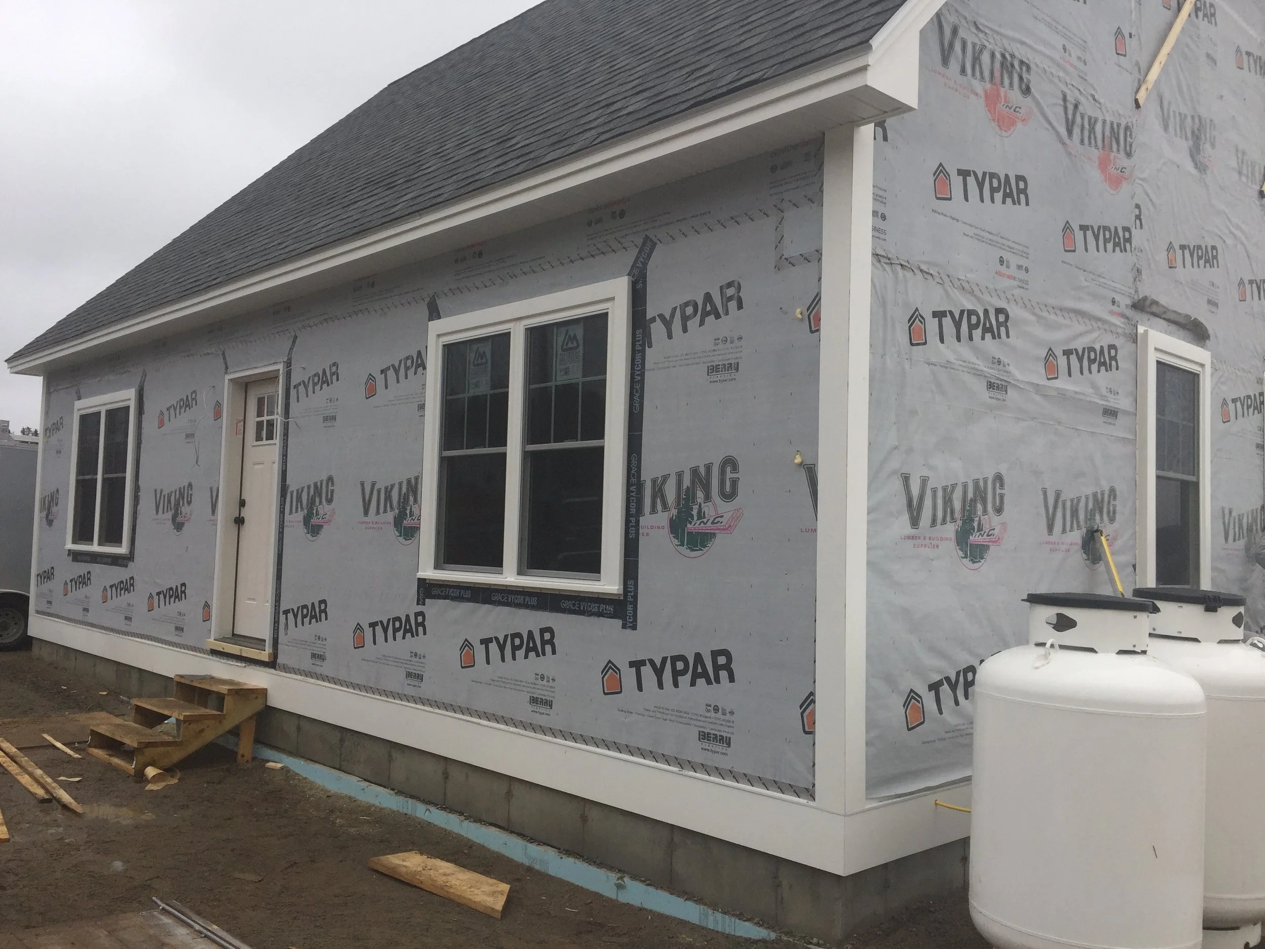 A house under construction with exterior walls covered in Typar weather-resistant barrier, two windows, and a door. There is a small set of wooden stairs leading to the door and construction materials on the ground, including a large white tank.